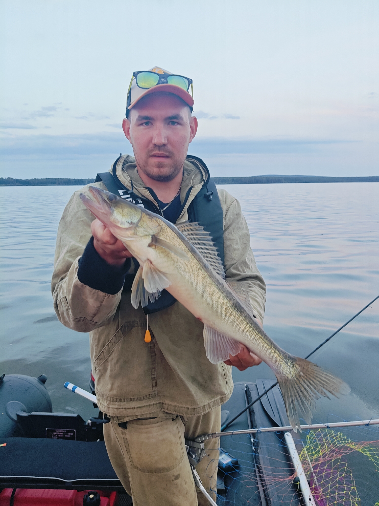 A man stands in a boat, holding a freshly caught fish. He is wearing a beige jacket, beige pants, and a cap with sunglasses on top. The fish, a walleye, is long and slender with a light green and silver body. The boat is on a calm body of water, with a tree-lined horizon in the background. The sky is overcast with some clouds. The scene appears to be from Chaykovsky, Russia, a city known for its water bodies and fishing opportunities.