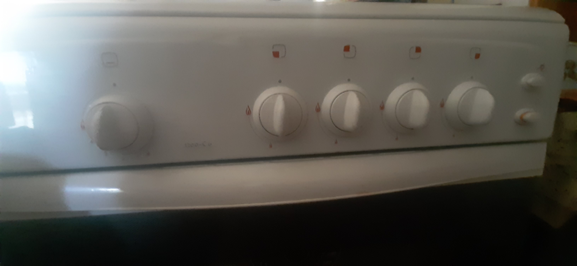 A close-up, eye-level shot shows the control panel of a white gas stove. The panel features four rotary knobs for controlling the burners, each with indicator marks. Above the knobs, there are symbols indicating burner settings. A model number, "1300-Cu," is visible below the third knob from the left. The stove is positioned indoors, likely in a kitchen in Yessentukskaya, Russia. The background is dark and out of focus, with a hint of a colorful, patterned object on the far right. The lighting suggests an indoor environment, with a bright reflection on the stove's surface.