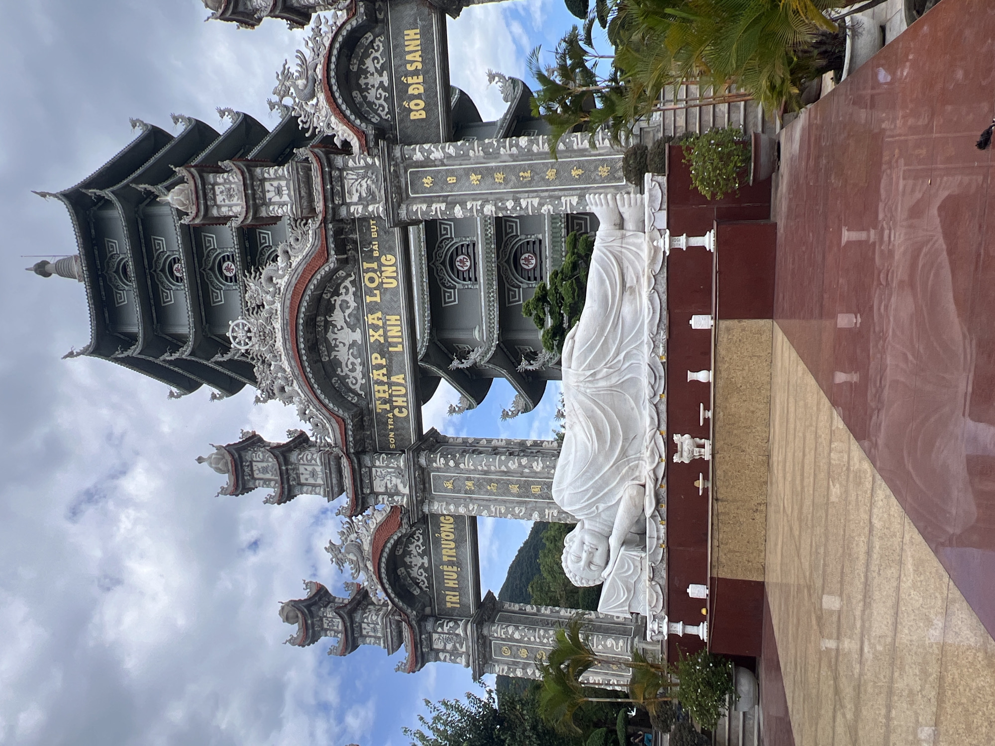 This image shows a large white marble statue of a reclining Buddha. The statue is lying on a raised platform in front of a multi-tiered, gray pagoda. The pagoda has ornate architectural details and is adorned with Chinese characters. The entire scene is set against a backdrop of a cloudy sky and lush green hills. The foreground features a polished floor, reflecting the colors of the surrounding structures and sky. The style of architecture and the presence of the reclining Buddha are characteristic of Buddhist temples in Vietnam. This particular statue is located at Linh Ung Pagoda on Son Tra Peninsula in Da Nang, Vietnam. There are no people clearly visible in the foreground or interacting with the statue. The environment appears to be a serene and spiritual setting, typical of a temple complex.
