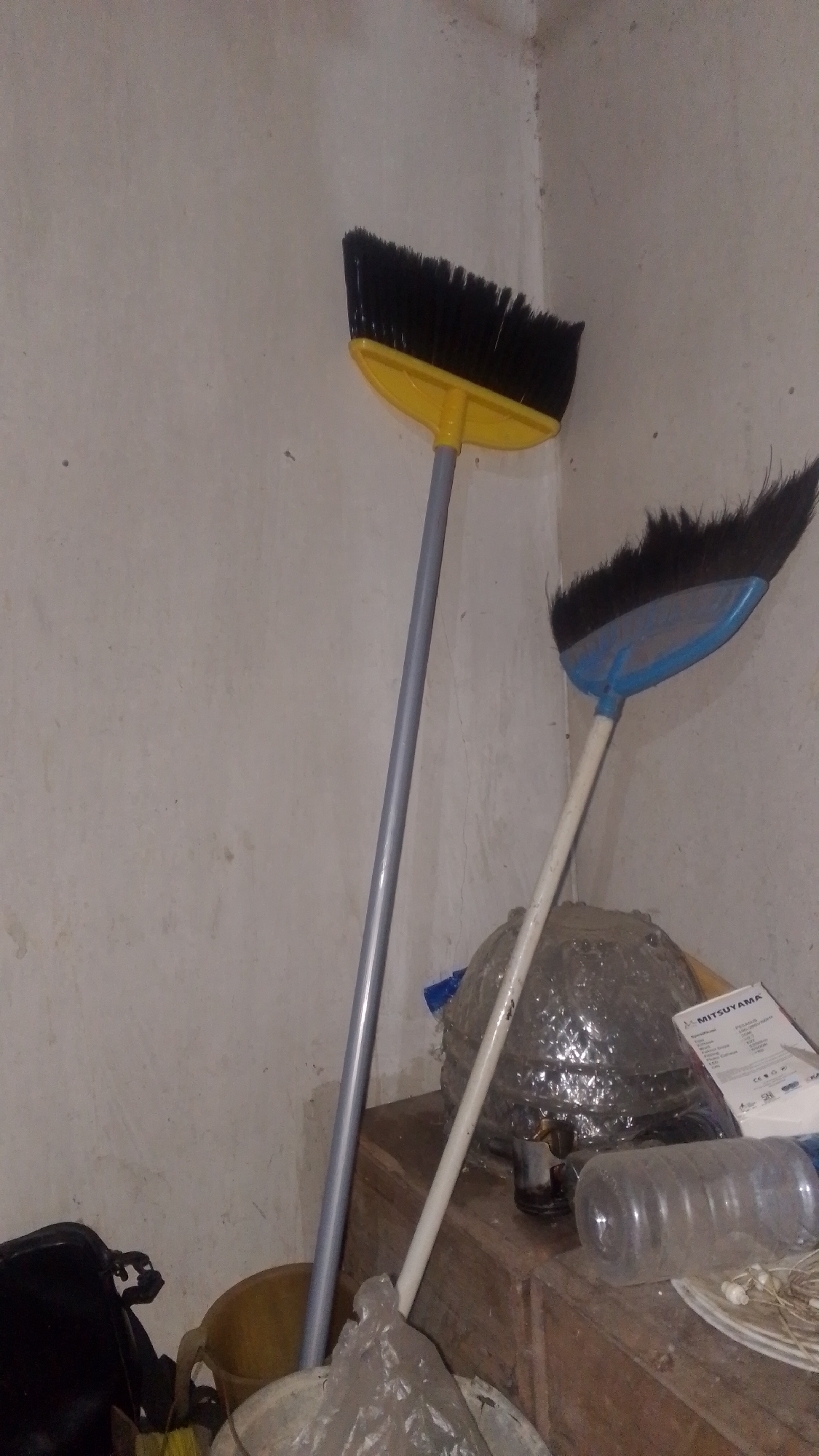 The image depicts a cluttered corner of a room, likely a storage area or a utility space, in Olak-Olak Kubu, Indonesia. Two brooms are prominently featured, standing upright and leaning against a textured, light-colored wall. One broom has a bright yellow plastic head and black bristles, with a long, silver-colored handle. The other broom has a blue plastic head and dark, possibly black or very dark brown, bristles, with a white handle.

In the foreground and midground, various objects are scattered. There is a large, roughly spherical object wrapped in clear plastic bubble wrap, situated behind the broom with the blue head. To the right of this, a wooden surface, possibly a table or cabinet, is visible. On this surface, a clear plastic water bottle is lying on its side, and a white box with the brand name "MITSUYAMA" printed on it is also present. In the lower left of the image, a black bag or sack is partially visible, along with a yellow bucket. Other unidentifiable items, including plastic bags and what appears to be some sort of wire or string, are also present in the lower portion of the frame.

The lighting in the image is dim, suggesting an indoor environment with limited natural light. The overall impression is one of disarray and a lack of organization. There are no people visible in the scene. The wall exhibits some discoloration and minor imperfections, consistent with an aging interior. The scene is forensic in its detail, documenting the objects and their arrangement without interpretation or emotional context.