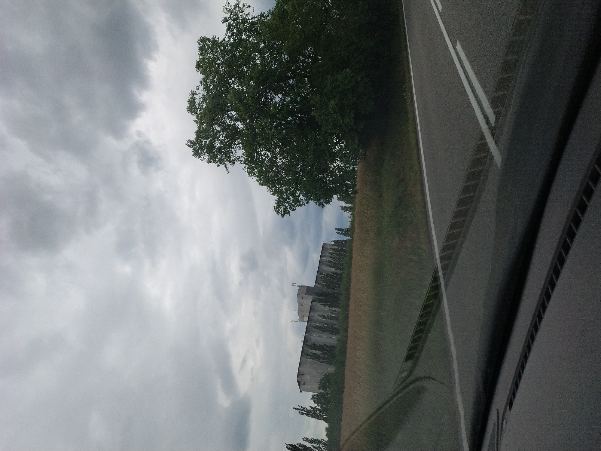 The image depicts a scene captured from a vehicle, likely from a dashboard camera, as evidenced by the reflection of the interior on the right side of the frame. The perspective is tilted, suggesting the vehicle is moving. The main subjects are a large, mature tree with dense green foliage and a long, multi-story building with a gray facade and numerous windows. The building appears industrial or agricultural, possibly a grain silo or warehouse, with antennas on its roof.
The setting is rural, with a paved road featuring white lane markings visible in the foreground on the right. To the left of the road, a grassy field and the aforementioned building are situated. Beyond the building, a line of trees stretches into the distance. The sky is overcast with heavy, gray clouds, indicating a cloudy or possibly stormy weather condition. The time of day is difficult to ascertain precisely due to the cloud cover, but it appears to be daytime.
The overall mood is somewhat somber and subdued, likely due to the overcast sky. No people are visible in the image. No text is discernible. The location context provided, Záhoří, Czechia, adds specific geographic information to the scene. The scene captures a snapshot of a journey through a rural landscape under a gray sky, with a prominent natural element (tree) and an industrial or utilitarian structure.