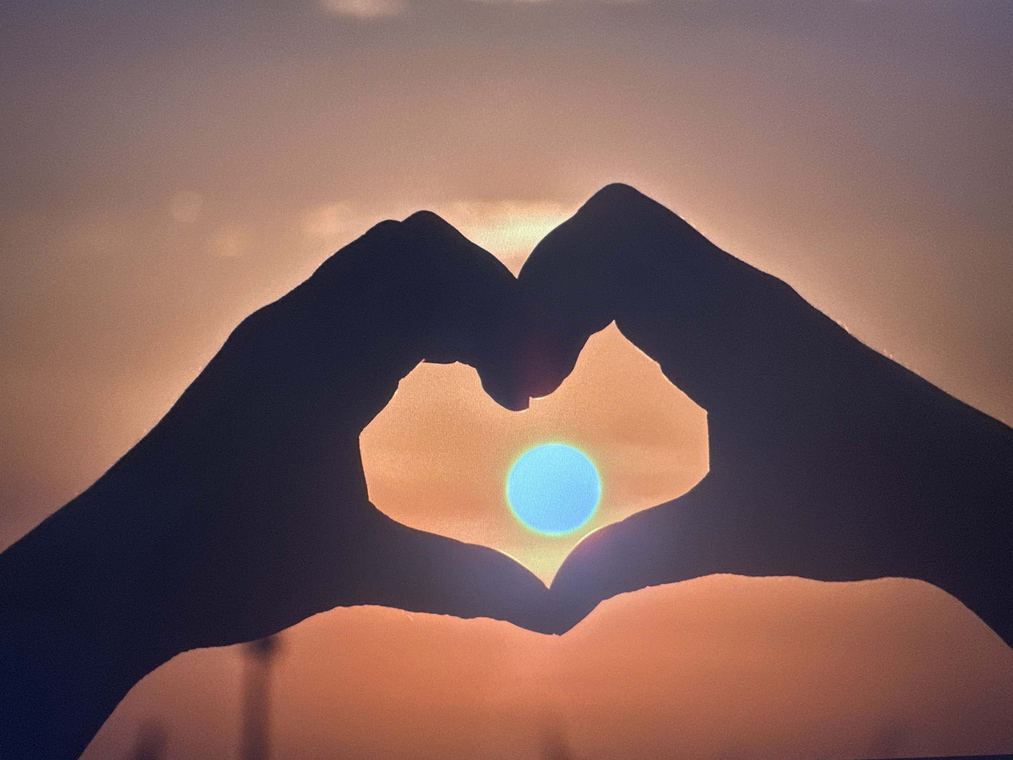 This is a silhouette of two hands forming a heart shape against a sunset. The sun, a bright blue circle, is positioned in the center of the heart. The sky is a gradient of orange and pink hues, with some faint clouds visible. The image does not provide any information about the city of Bryansk, Russia, or any specific location within it. There are no people, objects, or actions identifiable beyond the hands and the sun.