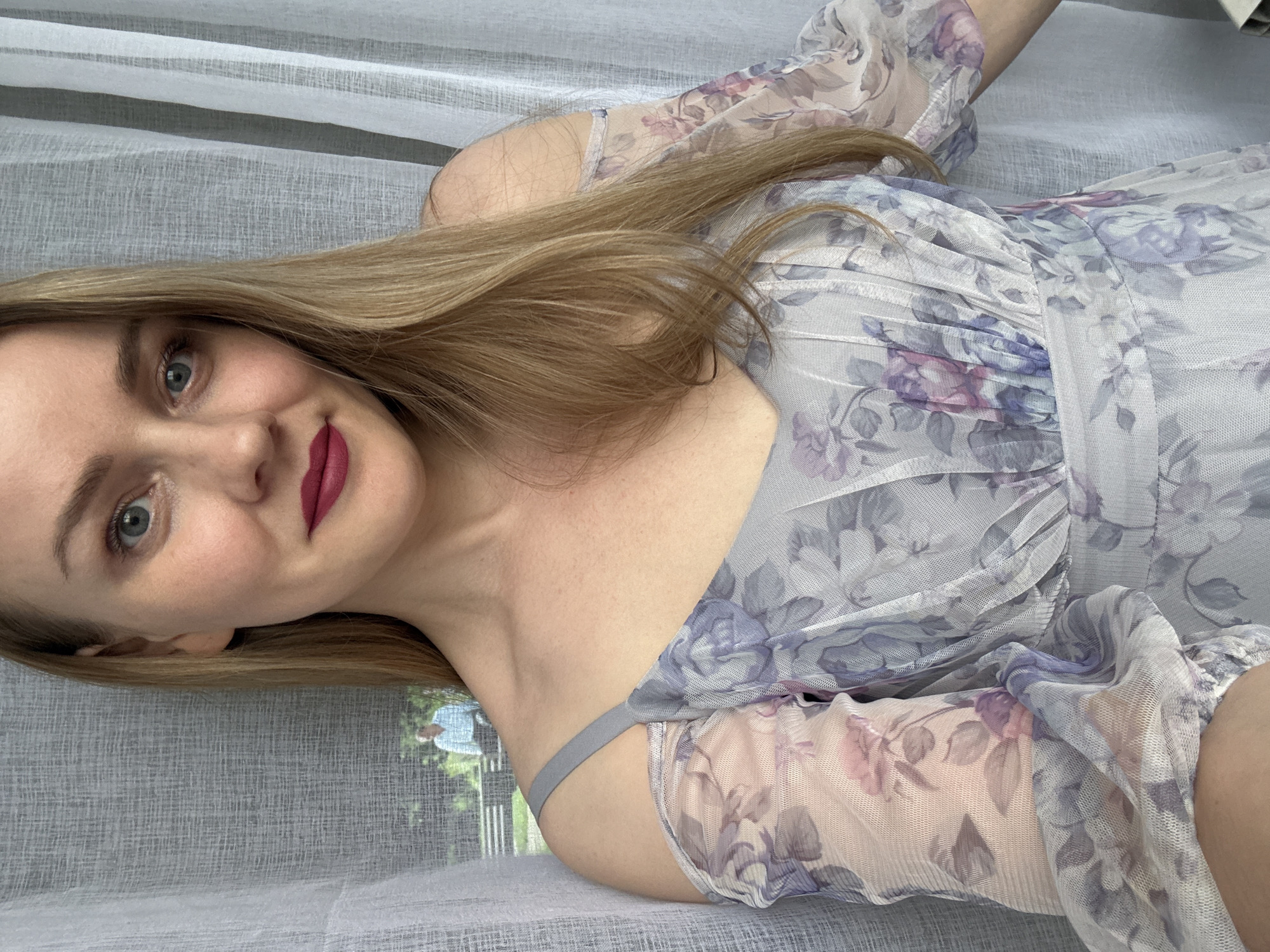 The image features a young adult woman with long, straight, light brown hair, lying on her side and looking directly at the camera with a gentle smile. She has light-colored eyes and is wearing dark pink lipstick. Her skin is fair.
She is dressed in a light-colored, sheer or mesh-like dress with a floral pattern in shades of purple and pink. The dress has long, somewhat puffy sleeves and a thin grey strap is visible on her left shoulder. The bodice appears gathered or ruched.
The setting is indoors, with the woman positioned against a backdrop of light, translucent curtains. Through a window, a blurred outdoor scene with greenery and hints of distant structures is visible. The surface she is resting on is covered in a similar light-colored, possibly textured fabric.
The activity taking place is a posed photograph, likely a selfie. The natural lighting suggests it is daytime. Her expression is calm and pleasant. There is no visible text in the image. The location is Chaikovsky, Russia.