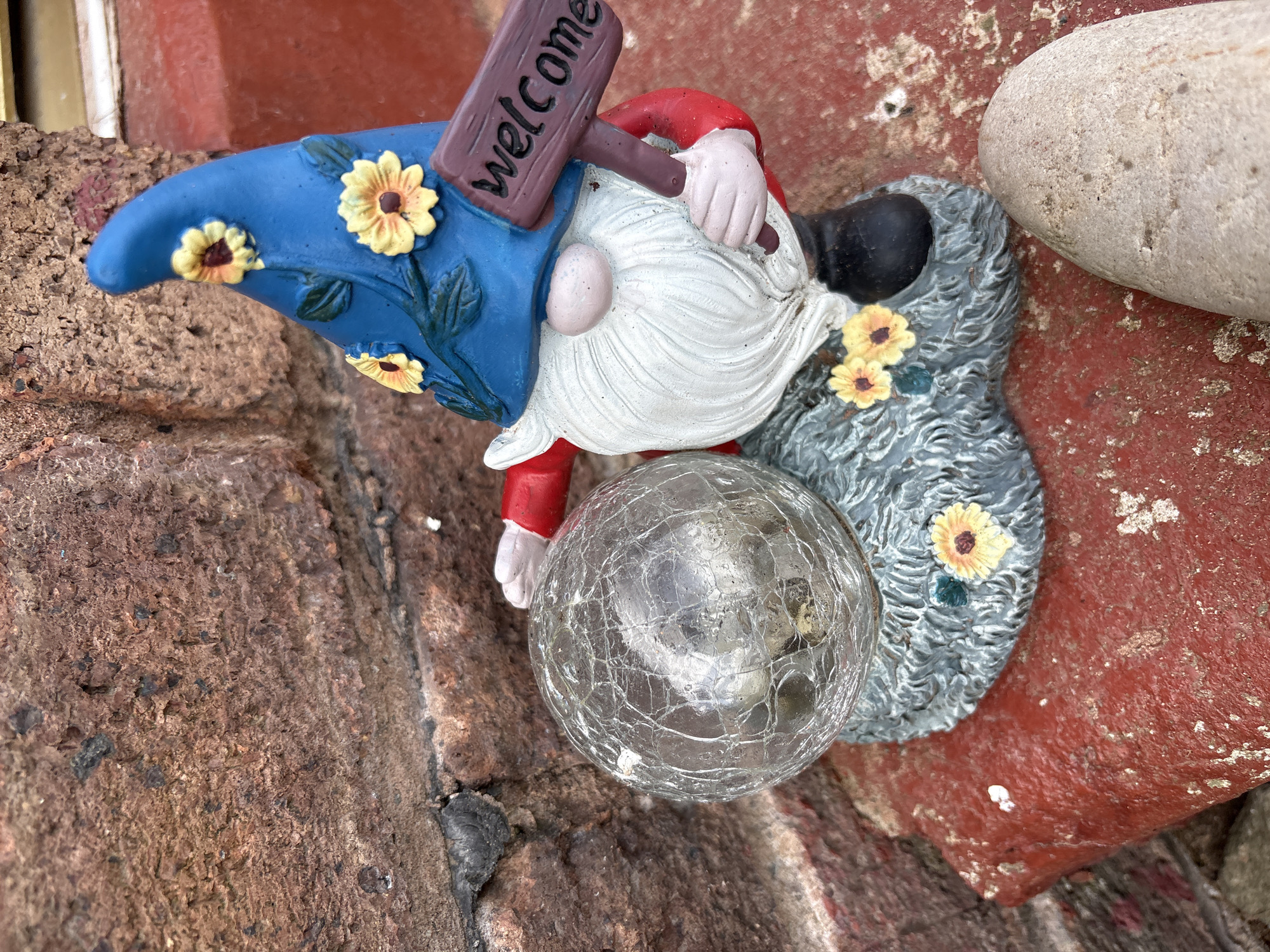 A close-up shot shows a garden gnome figurine on a rough, reddish-brown textured surface, likely brick or stone. The gnome has a blue hat decorated with yellow sunflowers, a white beard, and a red shirt. He holds a sign that reads "welcome". In front of the gnome is a cracked glass sphere. Beside the gnome is a smooth, light-colored stone. The setting suggests an outdoor location, possibly a garden or patio, and is likely located in Pulverbatch, United Kingdom.