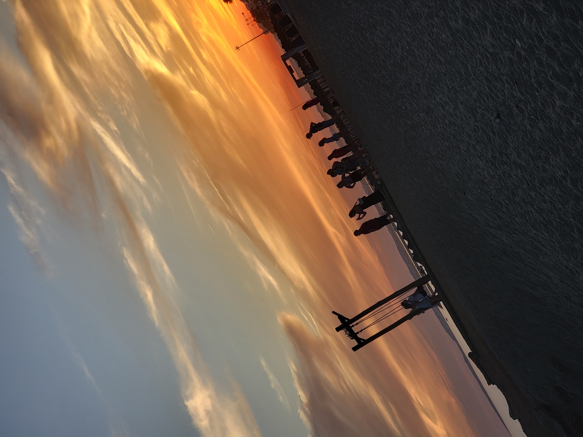 This image captures a beach scene in Quarteira, Portugal, during a vibrant sunset. The sky is dominated by hues of orange and yellow, with wispy clouds streaking across the expanse. In the foreground, a sandy beach is visible, with several people silhouetted against the bright sky. Some individuals are standing and looking towards the sea, while a group is gathered near a wooden swing structure. The sea is calm and reflects the colors of the sunset. In the background, buildings and palm trees are faintly discernible along the coastline, suggesting the urban edge of the beach. The overall atmosphere is peaceful and serene, characteristic of a beach at dusk.
