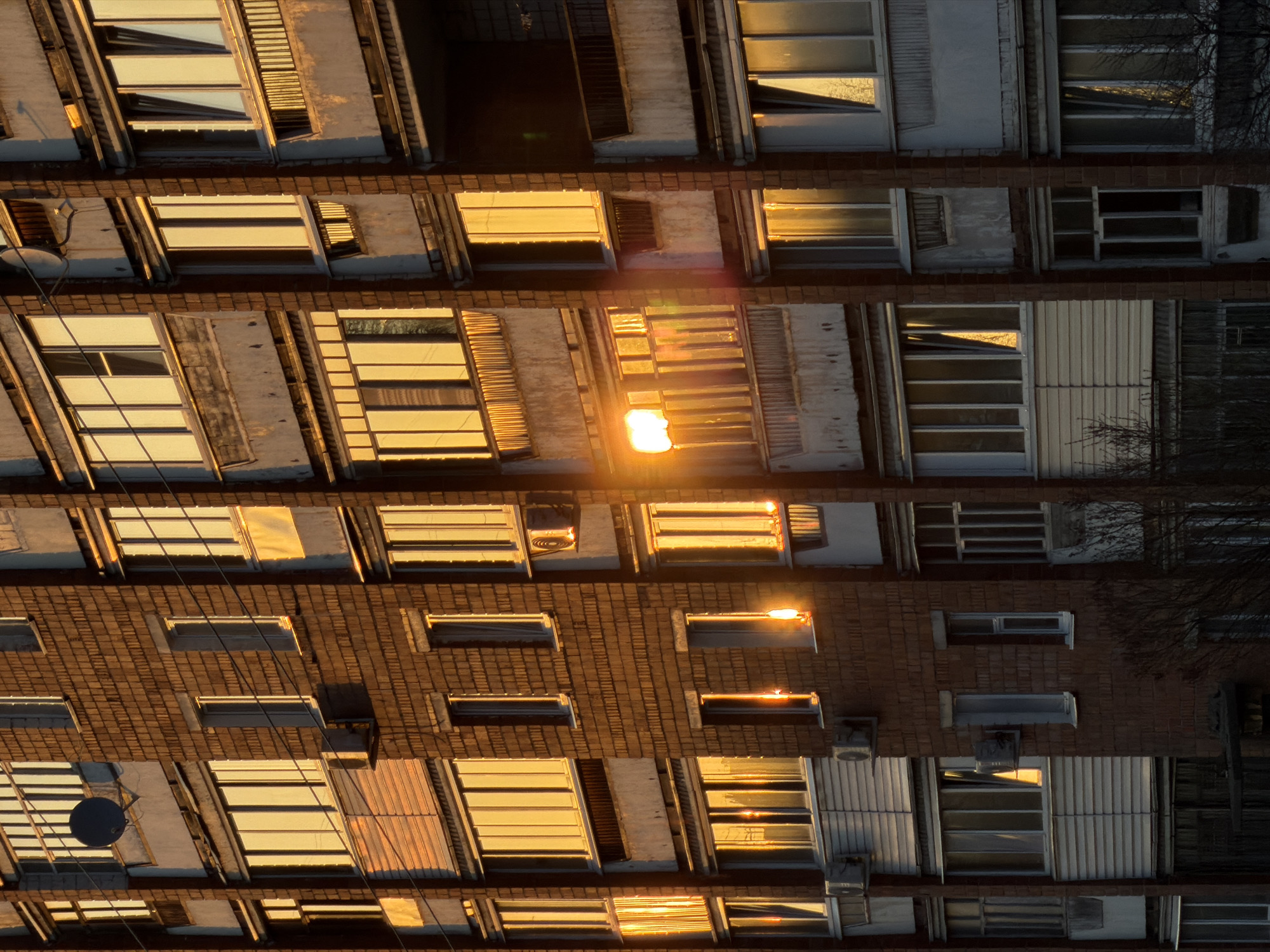 The image shows the facade of a residential building, likely located in Izhevsk, Russia, due to architectural similarities and material choices typical of Soviet-era construction in the city. The building is multi-storied, with a mixture of brick and prefabricated concrete panel construction. The windows reflect the warm tones of the setting sun, creating a bright, golden effect. Some windows are open, with visible blinds or curtains inside. The overall impression is that of an older, lived-in building, with visible signs of wear and tear on the facade. The photo is taken from below, pointing upwards at an angle, giving a slightly distorted perspective. There are no people visible in the image. The image focuses primarily on the architectural details and the interaction of light and shadow on the building's surface. Some satellite dishes are visible on the building's surface.
