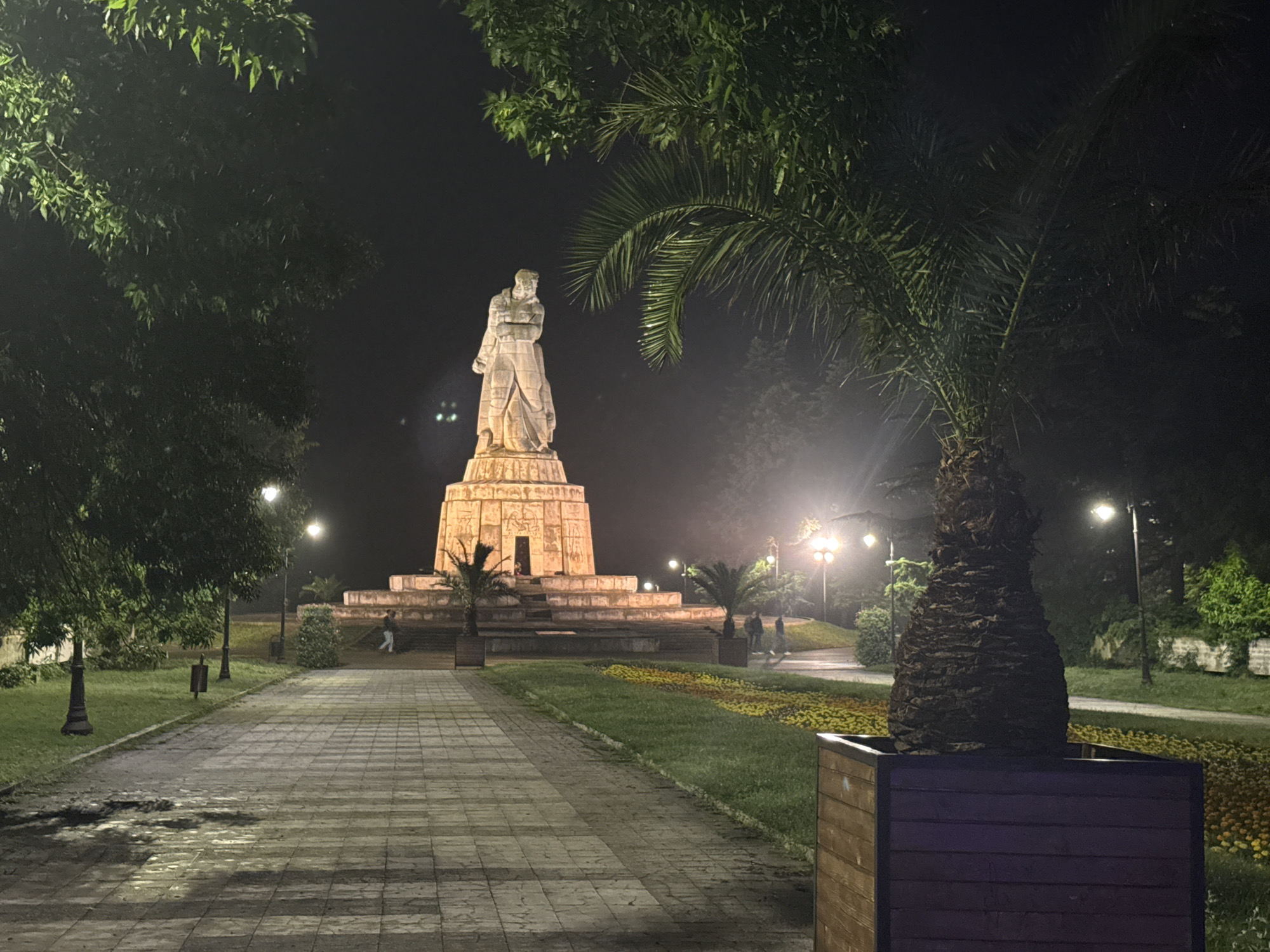 A nighttime view captures a brightly illuminated statue standing prominently on a multi-tiered pedestal in what appears to be a public park or plaza. The large, light-colored statue depicts a male figure in a long robe, with details carved into the pedestal. Spotlights from below cast a glow on the monument, making it a focal point against the dark sky.
Several people are visible walking in the mid-ground and background, near the base of the monument and along the pathways, suggesting a peaceful evening stroll. The scene is well-lit by numerous lampposts lining the paved pathways and illuminating grassy areas and trees. To the left, a paved path leads towards the monument, flanked by green grass and trees. On the right, a large palm tree in a wooden planter stands in the foreground, with other palm trees and various foliage scattered throughout the park. Flowerbeds with yellow and orange blooms are visible in front of the monument. The overall atmosphere is calm, with the artificial lighting highlighting architectural and natural features. No visible text is present in the image.
