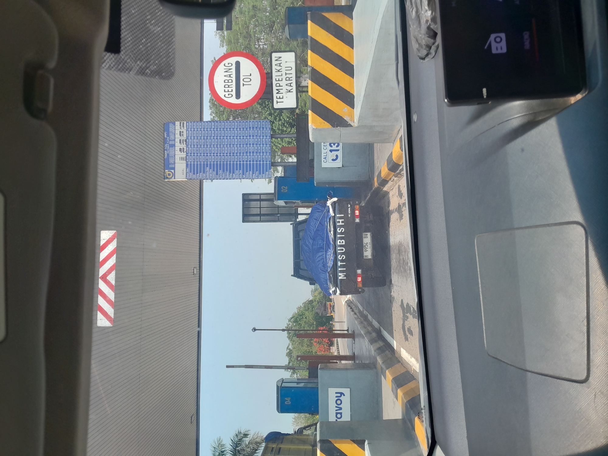 The image depicts a view from inside a vehicle approaching a toll booth in Kandeman, Indonesia. A black Mitsubishi pickup truck with a blue tarp covering its cargo is in the immediate foreground, stopped at the toll booth. The truck's license plate reads "H 9954 BV." To the left of the truck, a blue toll booth with the number "04" is visible, along with a sign that says "avoy." To the right of the truck, a circular red and white sign indicates "GERBANG TOL" (Toll Gate), and below it, a rectangular sign reads "TEMPELKAN KARTU" (Insert Card). Yellow and black striped barriers are present around the toll booth area. In the background, trees and a clear blue sky are visible. The scene is well-lit by daylight.