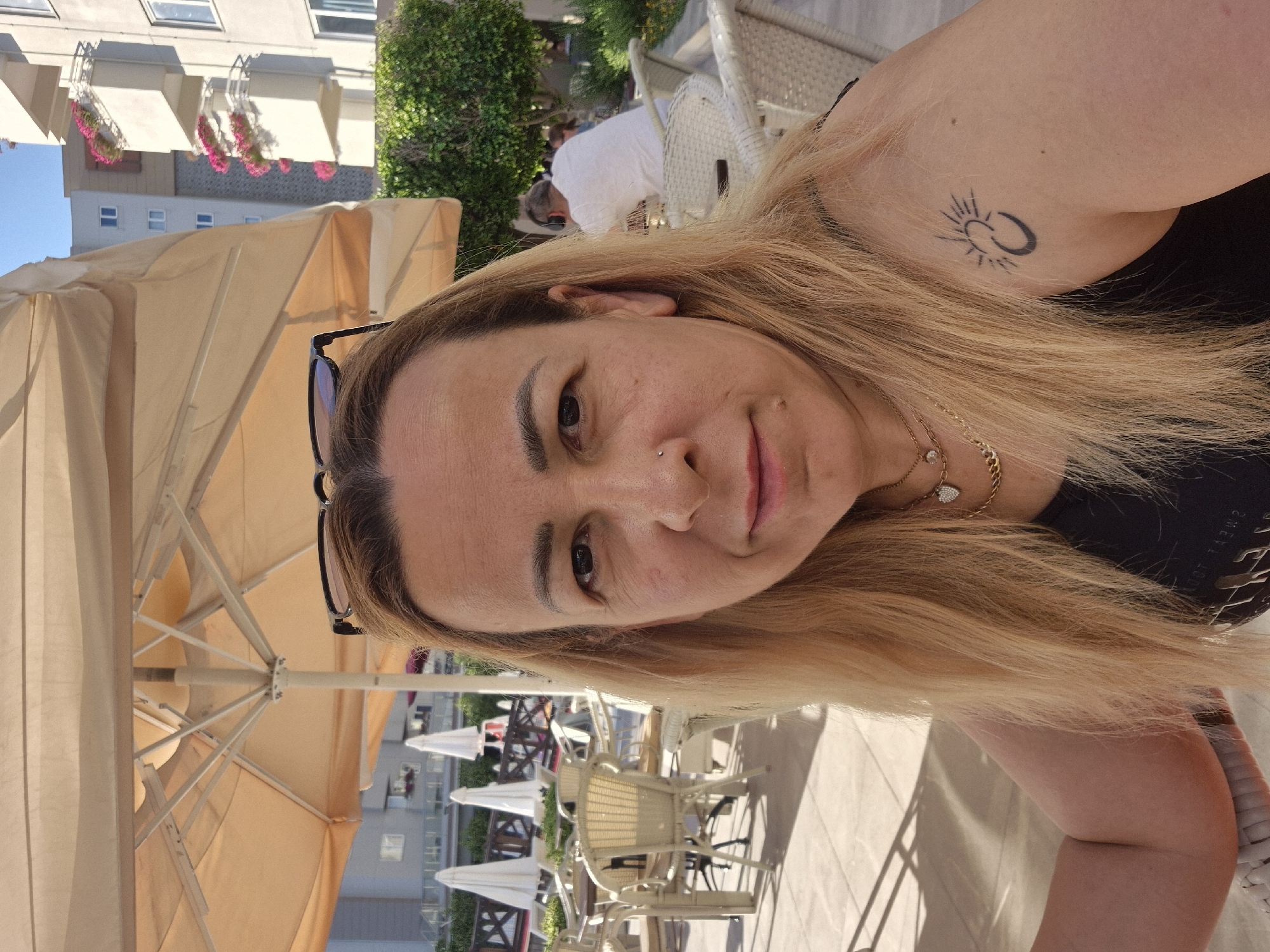 An outdoor, eye-level, medium shot captures a fair-skinned woman with shoulder-length blonde hair in a close-up selfie. She is wearing a black sleeveless top and several necklaces, one of which is gold with a heart-shaped pendant. A tattoo of a sun and crescent moon is visible on her left shoulder. Her head is tilted slightly to her left, and she is looking directly at the camera with a neutral expression. She wears black-rimmed glasses on top of her head. The background reveals a resort-like setting with white tables and chairs, beige umbrellas, and buildings with pink flowers on balconies, suggesting a warm-weather location. The lighting indicates it is daytime.