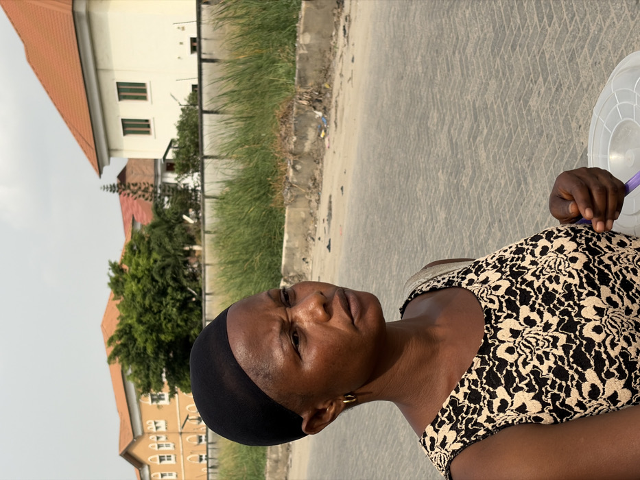 An adult female stands on a paved street in Igboefon, Nigeria, under an overcast sky. She is positioned in the left foreground, looking towards the right side of the frame with a serious expression, her brow slightly furrowed. She wears a black mesh wig cap, small gold hoop earrings, and a sleeveless top featuring an intricate black and beige/cream pattern. A light-colored strap, possibly from a bag, is visible over her right shoulder. Her right hand is seen in the lower right, holding a translucent white plastic container with a ridged lid, inside of which a purple utensil is partially visible. The paved street, composed of interlocking bricks, recedes into the background to the right. To the right of the street, a dirt verge containing scattered debris runs parallel to a low concrete barrier. Beyond the barrier, tall green grass grows. In the far background, several buildings with light-colored walls (yellow-orange and white) and prominent red-orange tiled roofs are visible, interspersed with lush green trees.