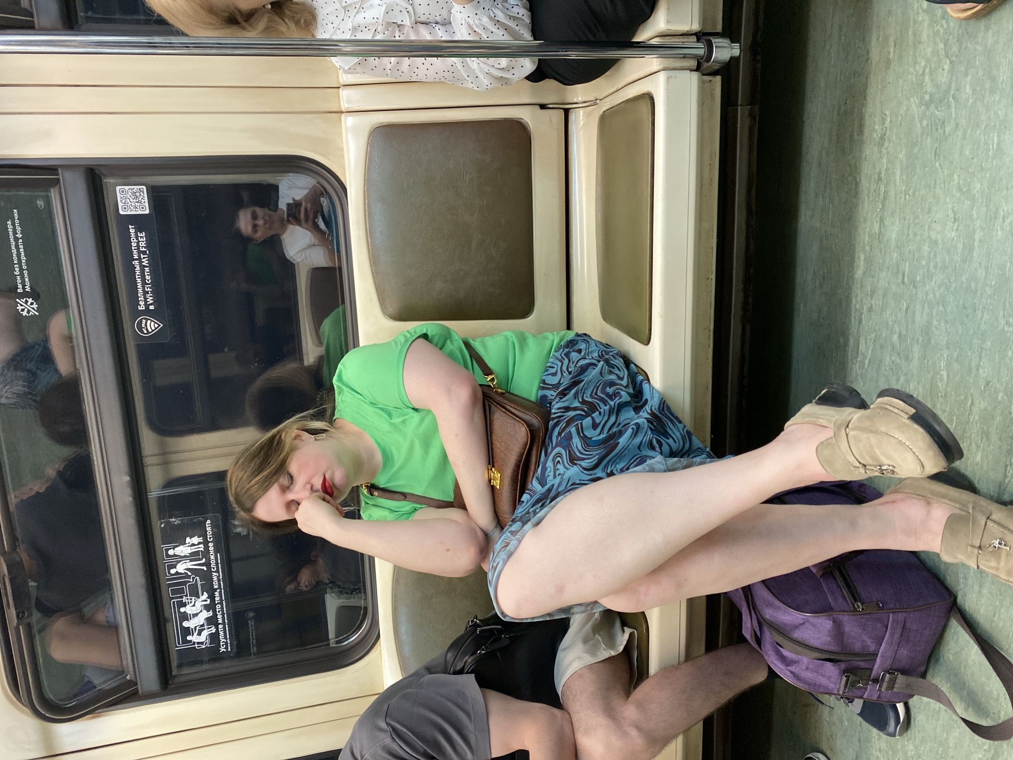 The image shows the interior of a train carriage in Moscow, Russia. A woman is sleeping, sprawled across a seat. She is wearing a green t-shirt, a blue patterned skirt, and beige loafers. A brown purse rests on her side, and her legs are stretched out on a purple backpack. Another person is seated next to her, wearing grey shorts. On the window next to the woman, there are stickers with pictograms indicating seating priorities for those with disabilities and a Wi-Fi free announcement. A reflection in the train window shows another man sitting. An older woman is also seen sitting in the background.