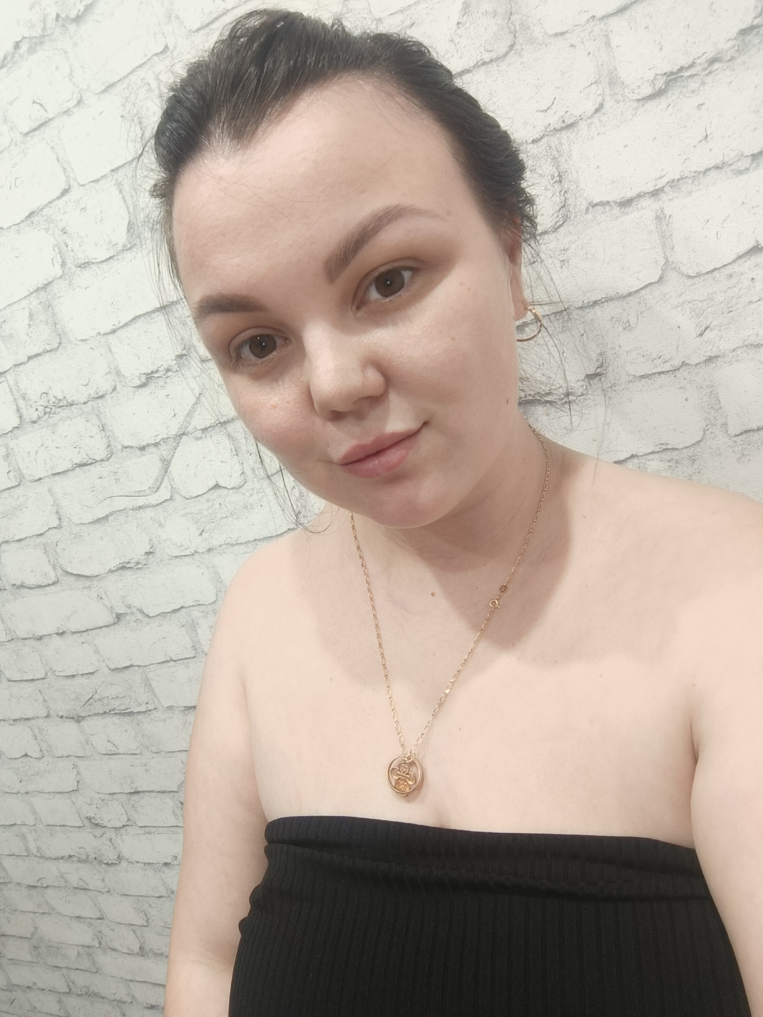 A selfie shows a woman with dark hair pulled back, wearing a black strapless top and a gold necklace with a circular pendant. She is facing the camera with a slight smile. The background is a white brick wall. The location is unknown.