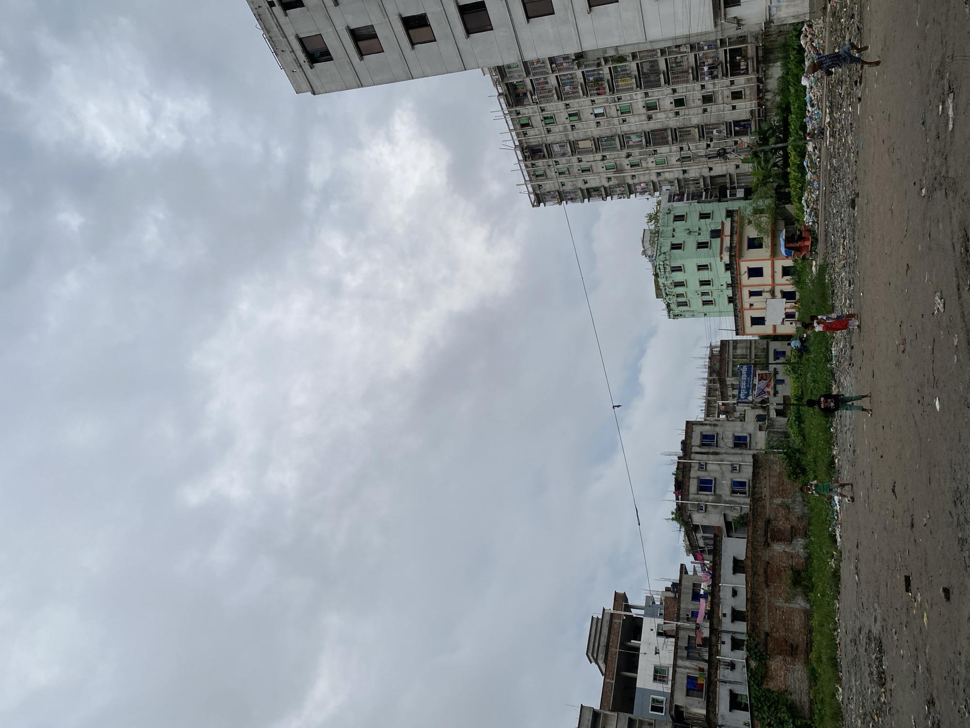 This is a vertical, eye-level, medium shot of an outdoor scene in Dhaka, Bangladesh, under a cloudy sky. In the foreground, two young boys are playing soccer on a dirt field littered with trash. One boy in a green shirt kicks a soccer ball, while the other in a black shirt stands watching. In the mid-ground, a row of buildings stretches across the frame. To the right, a tall, modern apartment building dominates the scene, its white facade punctuated by numerous dark windows. Next to it is a gray, multi-story building with a complex facade of balconies and windows. To the left of these, a smaller, pale green building with orange trim stands out, followed by a series of older, more dilapidated buildings. Clothes are hung out to dry on some of these structures. In the background, more buildings, some appearing to be under construction, are visible. A small red auto-rickshaw is parked near the green building, and piles of trash are scattered throughout the mid-ground and foreground. The overall impression is one of urban density and contrasting development.