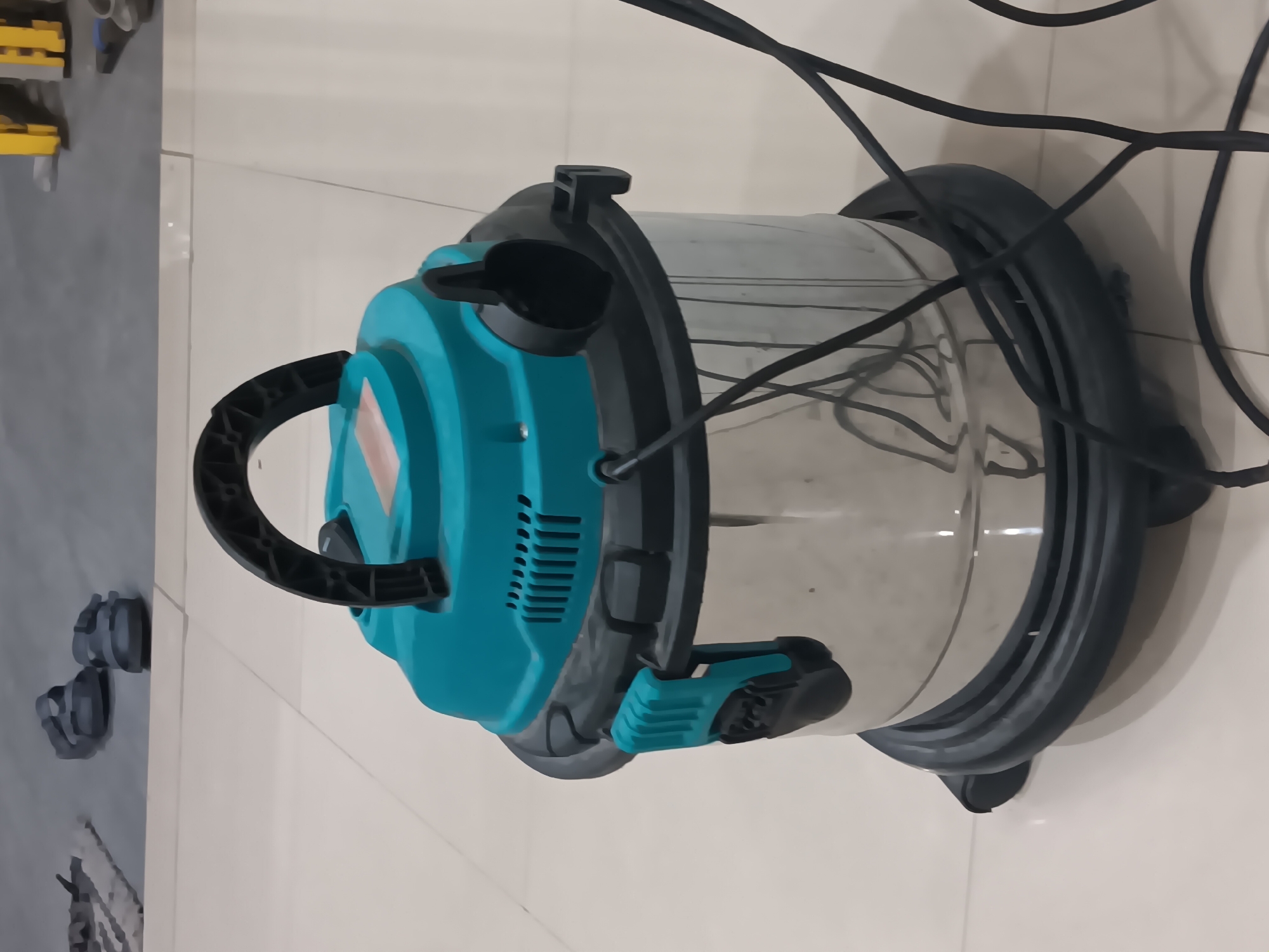 A teal and silver wet/dry vacuum cleaner sits on a tiled floor in what appears to be a residential or workshop setting in Batursari, Indonesia. The vacuum has a black handle on top and a clear plastic hose connector. Black power cords are visible on the floor around the vacuum. In the background, there are several pairs of sandals on a gray tiled surface, and a yellow industrial shelving unit. The lighting suggests an indoor environment with artificial light.