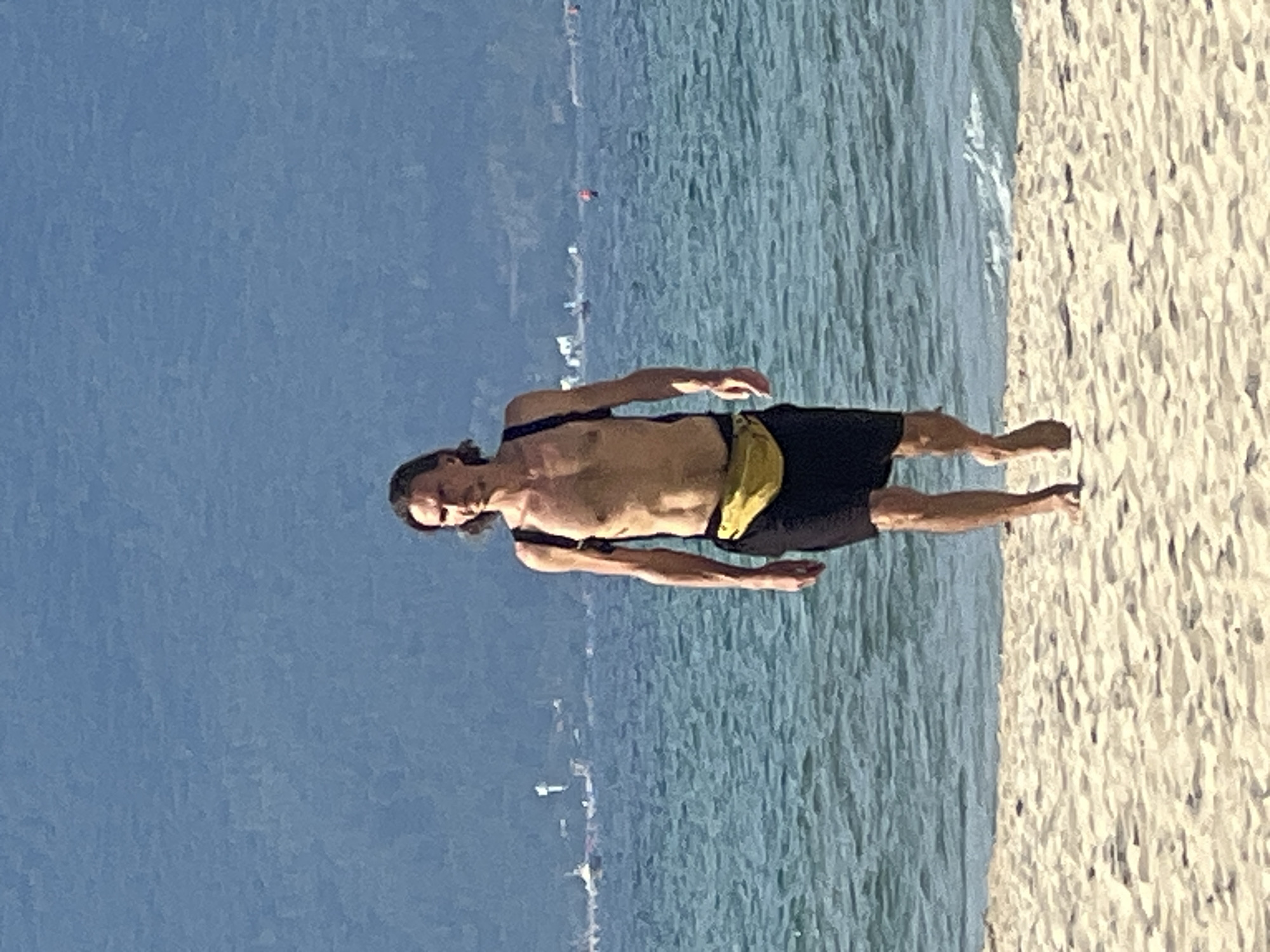 A man with long brown hair, shirtless and wearing black shorts and a yellow fanny pack, walks on a sandy beach towards the viewer. He is standing near the water's edge, with the ocean and a hazy blue mountain range in the background. Several boats are visible on the water. The image appears to have been taken in Da Nang, Vietnam.