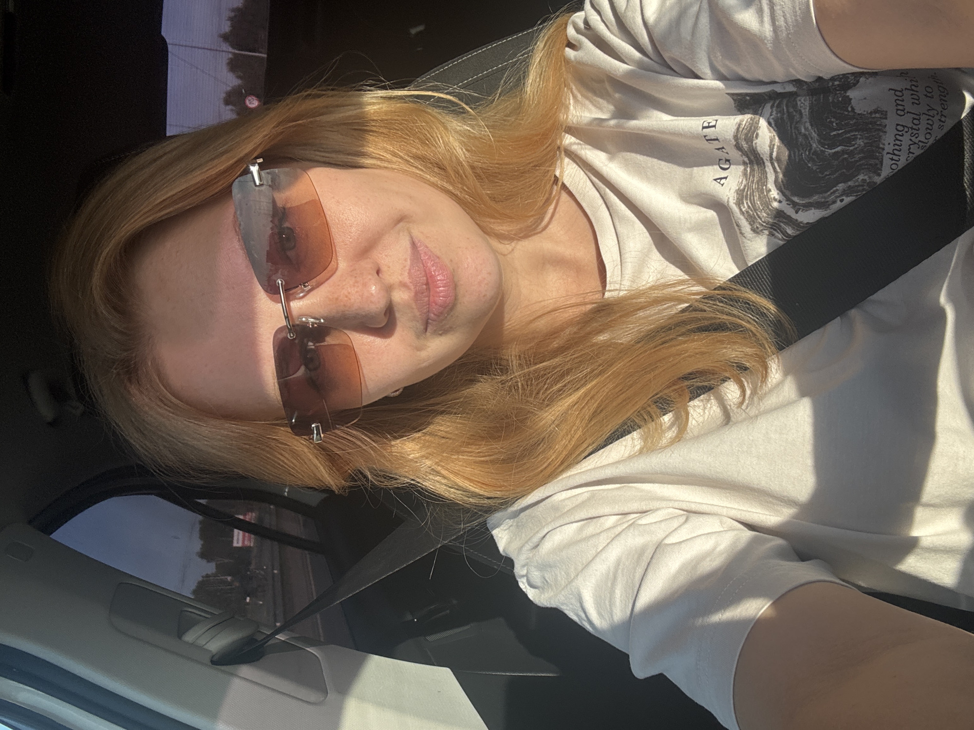 A young woman with shoulder-length blonde hair and fair skin is sitting in the passenger seat of a car. She is wearing sunglasses with orange lenses and a white t-shirt with a graphic print. The t-shirt has the word "AGATE" printed at the top and an image of a stone or mineral below it, along with text that reads "othing and crystal wh slowly to strength." The seatbelt is visible across her chest. The car is moving, as indicated by the blurred scenery outside the window, which includes trees and a road. The sun is shining, casting shadows on the woman and the interior of the car. The overall impression is a candid snapshot taken during a daytime drive. The image's metadata indicates the location as Zhukovo, Russia.