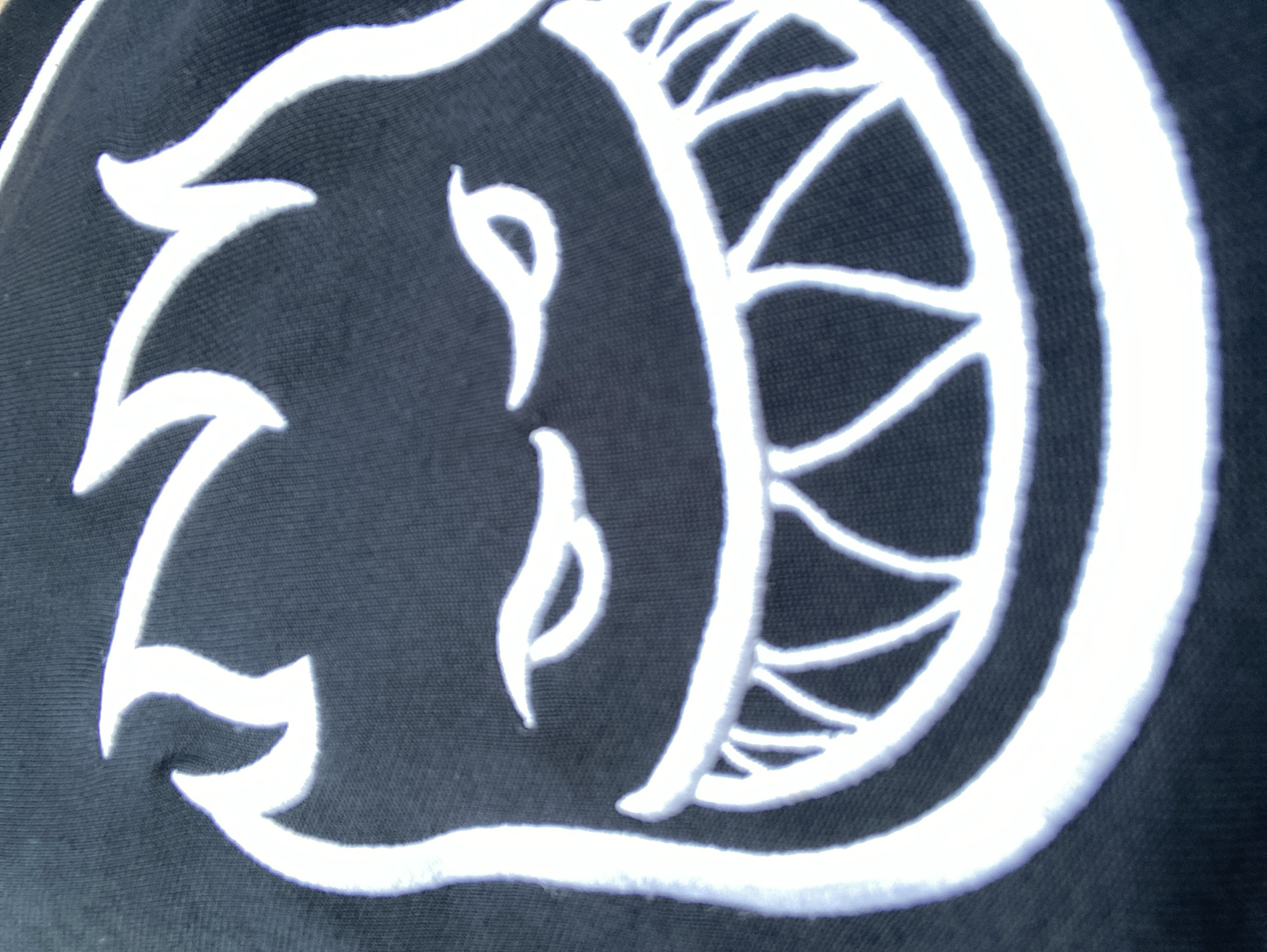 The image shows a close-up, detailed view of a white embroidered graphic of a stylized flaming skull with sharp teeth on a dark fabric, likely apparel. The embroidery is thick and textured, with a slight sheen reflecting the light. The skull's features are defined by thick white stitching, creating bold outlines for the flames, eyes, and mouth. The eyes are narrow and slanted, conveying an aggressive or mischievous expression. The mouth is wide open, revealing numerous sharp, triangular teeth. The fabric appears to be a dark knit material, possibly cotton, with a visible weave pattern. There are no people or environmental elements present; the focus is entirely on the embroidered design. The location in Barcelona, Spain is not determinable from the visual information provided.