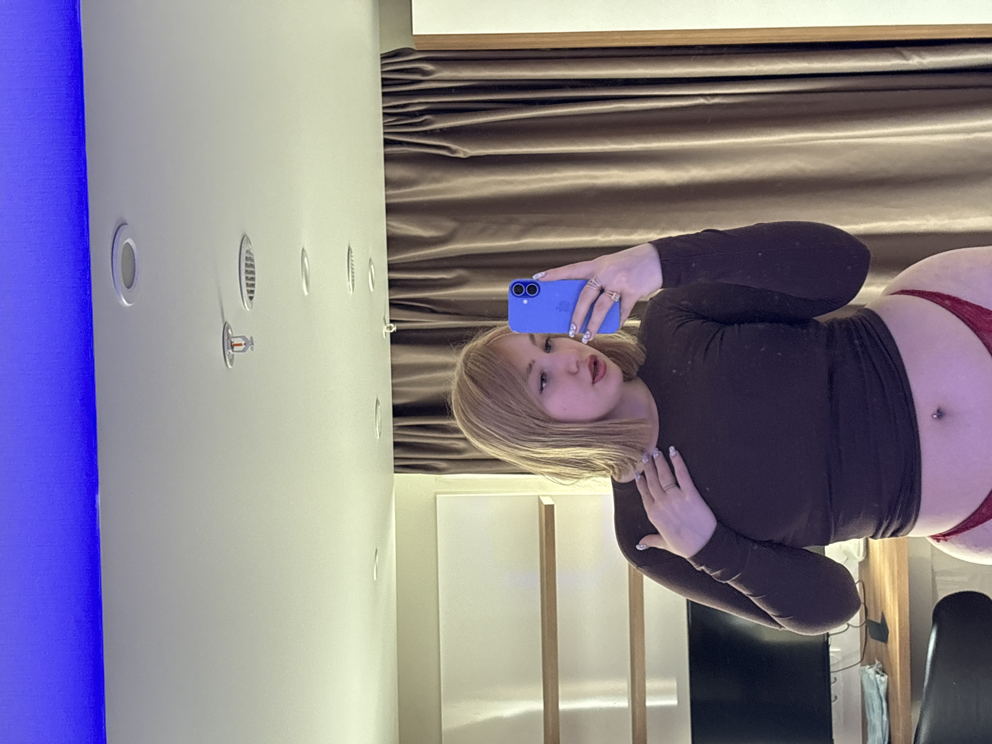 A young woman with blonde hair is taking a selfie in a room with beige curtains and blue lighting. She is wearing a dark brown long-sleeved shirt and red underwear. She is holding a blue smartphone in her right hand and has her left hand on her chest. The room appears to be a hotel room, as suggested by the modern decor and the presence of a computer monitor and shelves. The city of Khimki, Russia is known for its modern architecture and growing business district, which aligns with the visual cues in the image.