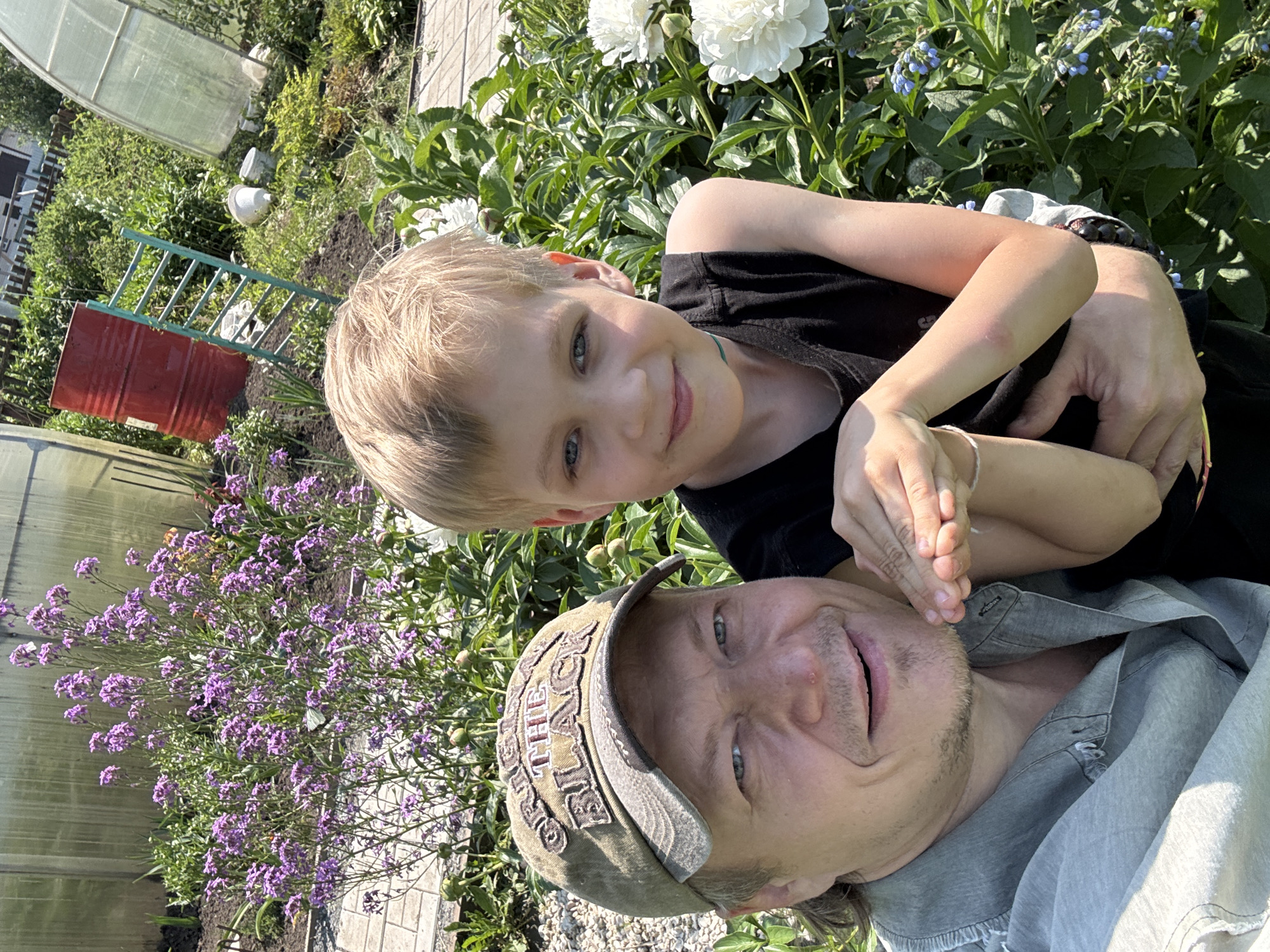 This is a selfie taken outdoors in a garden. The subjects are a boy and a man, likely father and son, who are posing for the camera. The boy, with blonde hair, is positioned in the foreground, looking directly at the camera with a slight smile. He is wearing a black t-shirt. The man, wearing a camouflage baseball cap and a light grey shirt, is in the background, also looking at the camera with an open-mouthed smile.
The setting is a lush garden with various plants, including large white peonies and smaller purple flowers. The background also shows hints of a greenhouse, a red barrel, and a green ladder, suggesting a rural or suburban environment. The sunlight indicates it is daytime, and the presence of flowers suggests it is likely spring or summer. The overall mood appears happy and relaxed, with the two individuals sharing a close moment.
There is visible text on the man's baseball cap, which appears to be embroidered letters, but they are not clearly legible. The image is oriented vertically, with the heads of the subjects at the top and bottom of the frame respectively.