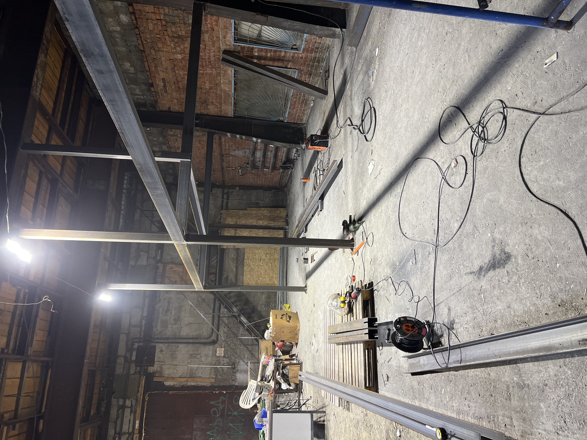 The image depicts the interior of a building under construction in an unknown location. A steel framework has been erected, dominating the central view. A brick wall with barred windows is visible in the background. The concrete floor is cluttered with various tools and materials, including a welding machine, power cords, metal beams, a pallet, a table, and a chair. In the lower portion of the image, a white screen and a box are located near what appears to be a utility cart. The lighting is a mixture of natural light from the window and artificial light from work lights, creating a stark contrast. The environment is industrial and active, indicative of ongoing construction. There are no people visible in the image.
