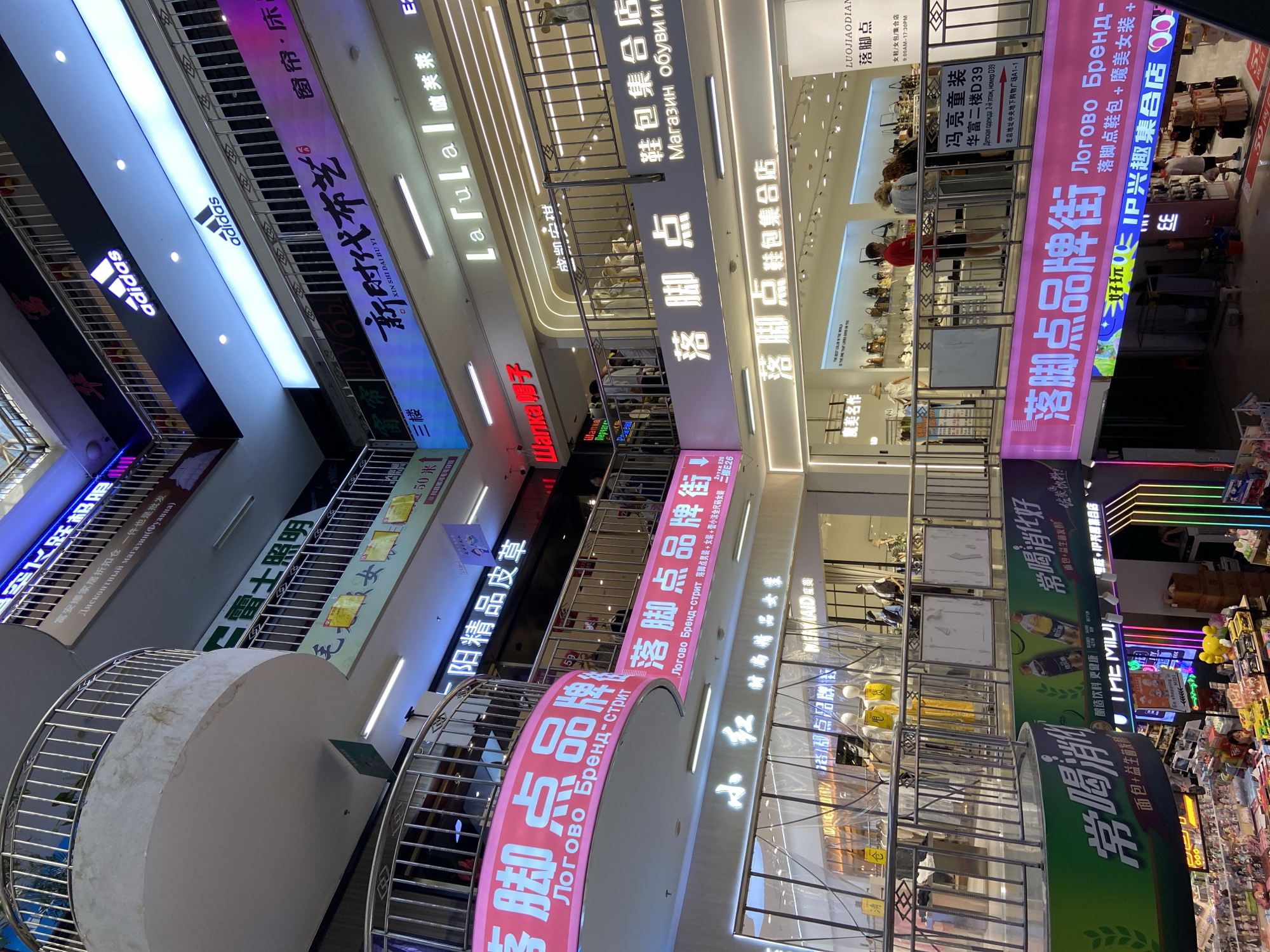 This is an interior shot of a multi-level shopping mall in Xingan Subdistrict, China. The mall features a modern design with escalators and balconies. Several shops are visible, with prominent signage in both Chinese and Russian. One large pink banner displays the text "落脚点品牌街" (Luòjiǎo diǎn pǐnpái jiē) which translates to "Luojiao Brand Street," along with Russian text that appears to be a translation. Another banner advertises "鞋包集合店" (Xié bāo jíhé diàn), meaning "shoe and bag collection store." There are also prominent Adidas logos visible on higher levels. People are seen browsing in some of the shops. The overall environment is brightly lit with a mix of neon and white lighting, showcasing a variety of retail offerings.