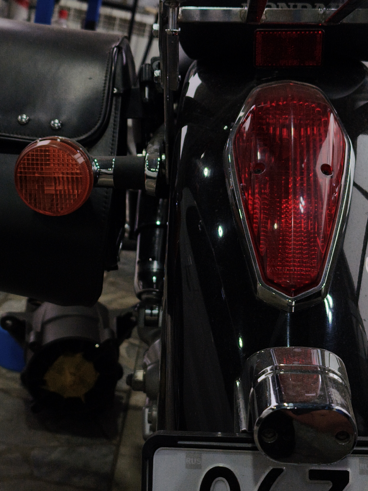 The image is a close-up, medium-shot photograph of the rear of a black motorcycle. The focus is on the taillight, a large, red, V-shaped assembly with a chrome bezel. To the left of the taillight, a portion of a black leather saddlebag is visible, with decorative metal studs. A circular orange reflector or turn signal is also on the left side. The motorcycle appears to be parked on a grey, textured surface, possibly concrete or asphalt. The background is out of focus but suggests an indoor environment with some metal structures. There are no people visible in the image. The photograph is taken from a low angle, looking upwards at the motorcycle. There is no information in the image to indicate its location in Noviy, Russia.