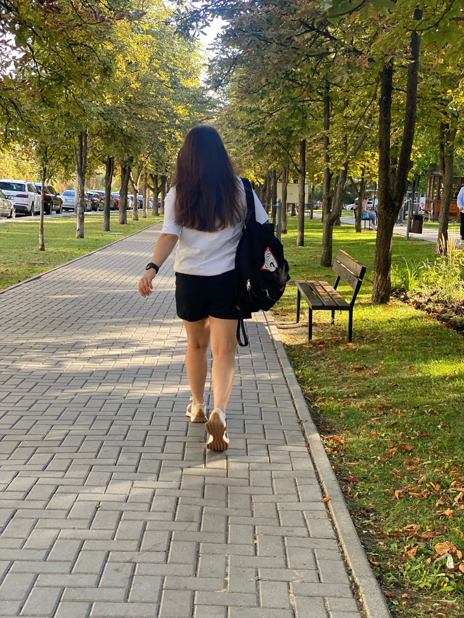 A young woman with long, dark hair is walking away from the camera on a paved pathway in a park. She is wearing a white t-shirt, black shorts, and light-colored sandals. She has a black backpack slung over her left shoulder. The pathway is made of gray interlocking pavers and is lined by trees with green and yellowing leaves, suggesting it is autumn. To the left of the pathway, there is a grassy area with parked cars visible in the distance. To the right of the pathway, there is more grass, a wooden park bench, and more trees. The sun is shining, casting shadows on the pathway and illuminating the leaves of the trees. The scene appears to be in a public park in Chișinău, Moldova, possibly near a street given the presence of parked cars.