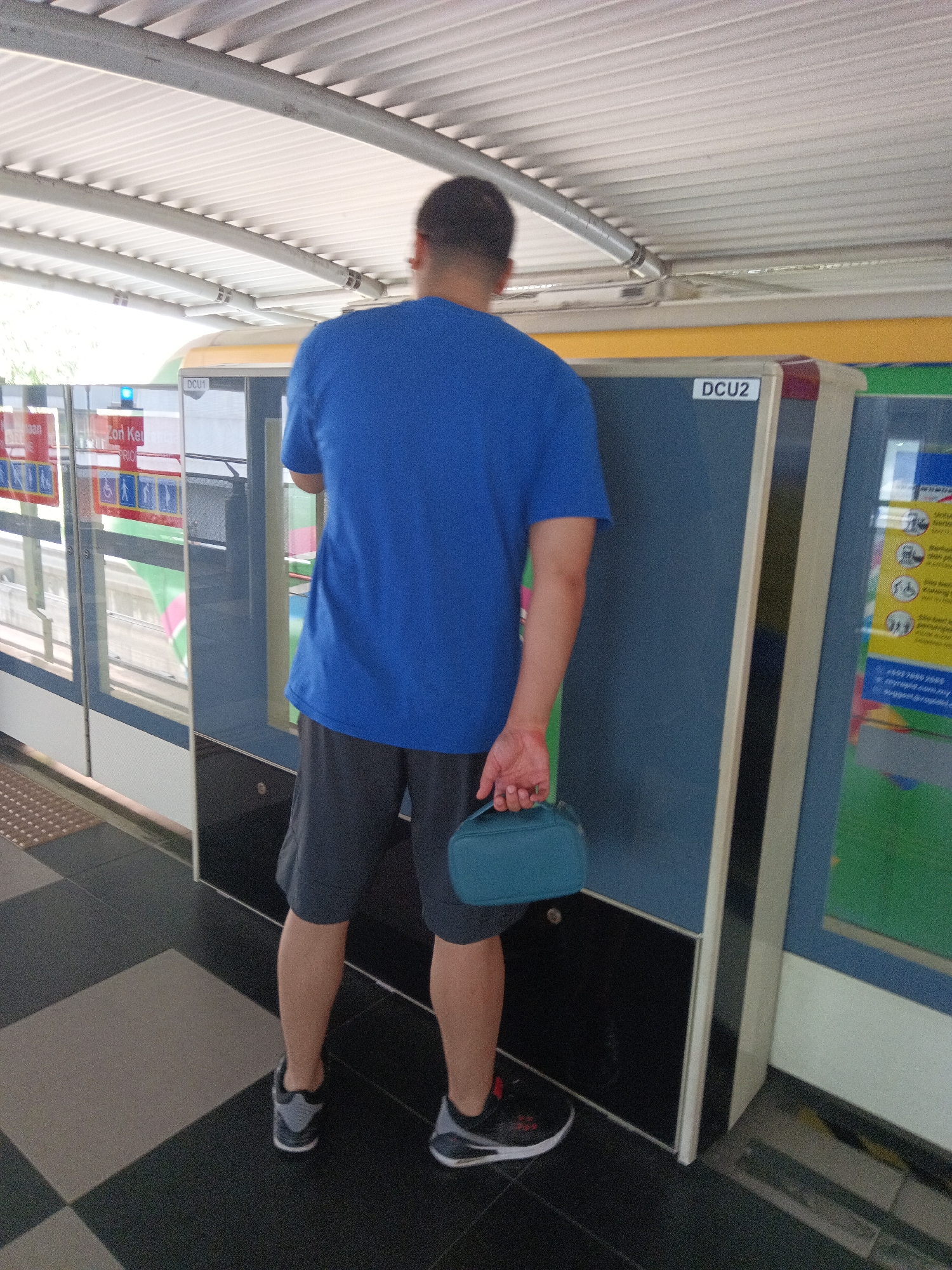 A man, viewed from behind, stands on a checkered tile floor in what appears to be a covered transit station. He is wearing a blue t-shirt and dark shorts, with black and grey athletic shoes. In his right hand, he holds a small, teal-colored zippered pouch. To his left, a train car with reflective windows is visible, marked with signs in what looks like Malay. To his right, there are two vertical panels, one blue and one green, with the label "DCU2" on the blue panel. A yellow and green sign with pictograms and text in Malay is attached to the green panel. The ceiling of the station is made of corrugated metal panels supported by metal beams.