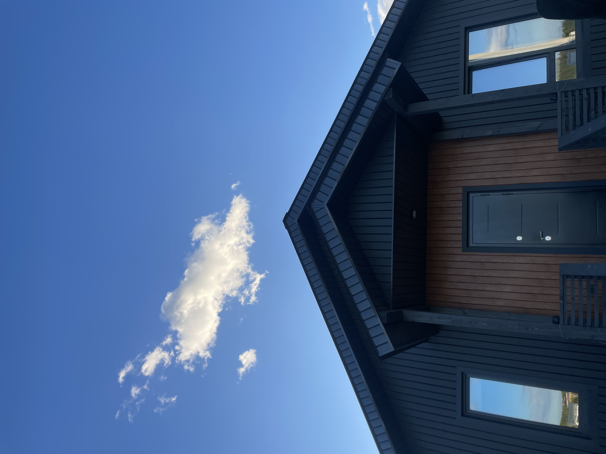 A low-angle shot captures the corner of a building against a vibrant blue sky with a single cumulus cloud. The building is a two-story structure with a dark exterior, contrasted by a section of wood paneling. A dark door and two windows with visible reflections add detail to the facade. The location is potentially Подшивалово, Russia, but without further context, this is speculative.