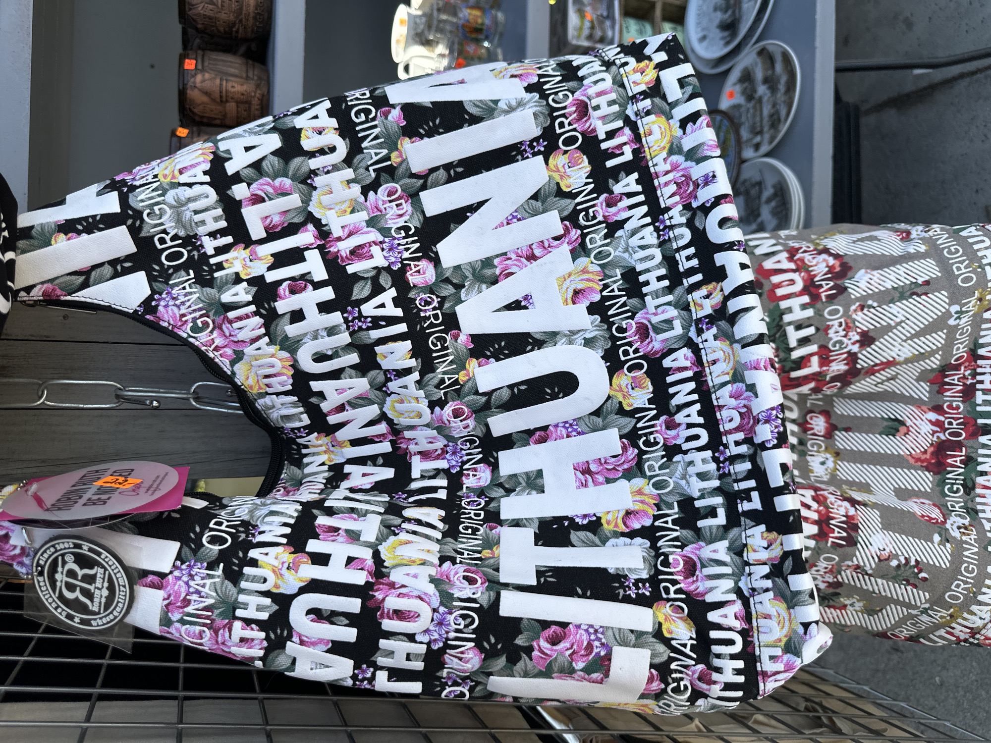 A close-up, eye-level, outdoor, medium shot captures a black fabric tote bag adorned with a floral pattern and repeated white text reading "LITHUANIA" in large block letters and "ORIGINAL" in smaller script. The bag is hanging by a silver chain, partially obscuring a wooden plank background and a wire fence to the left. A pink tag with yellow price stickers and a circular tag with "Robin Ruth" and an "RR" logo are attached to the bag's strap. In the blurred background, shelves display what appear to be ceramic mugs and plates. The scene is likely an outdoor market or souvenir shop in Trakai, Lithuania, given the bag's prominent "LITHUANIA" text and the general aesthetic of such locations. The focus is on the bag and its design, with the surrounding environment providing context.
