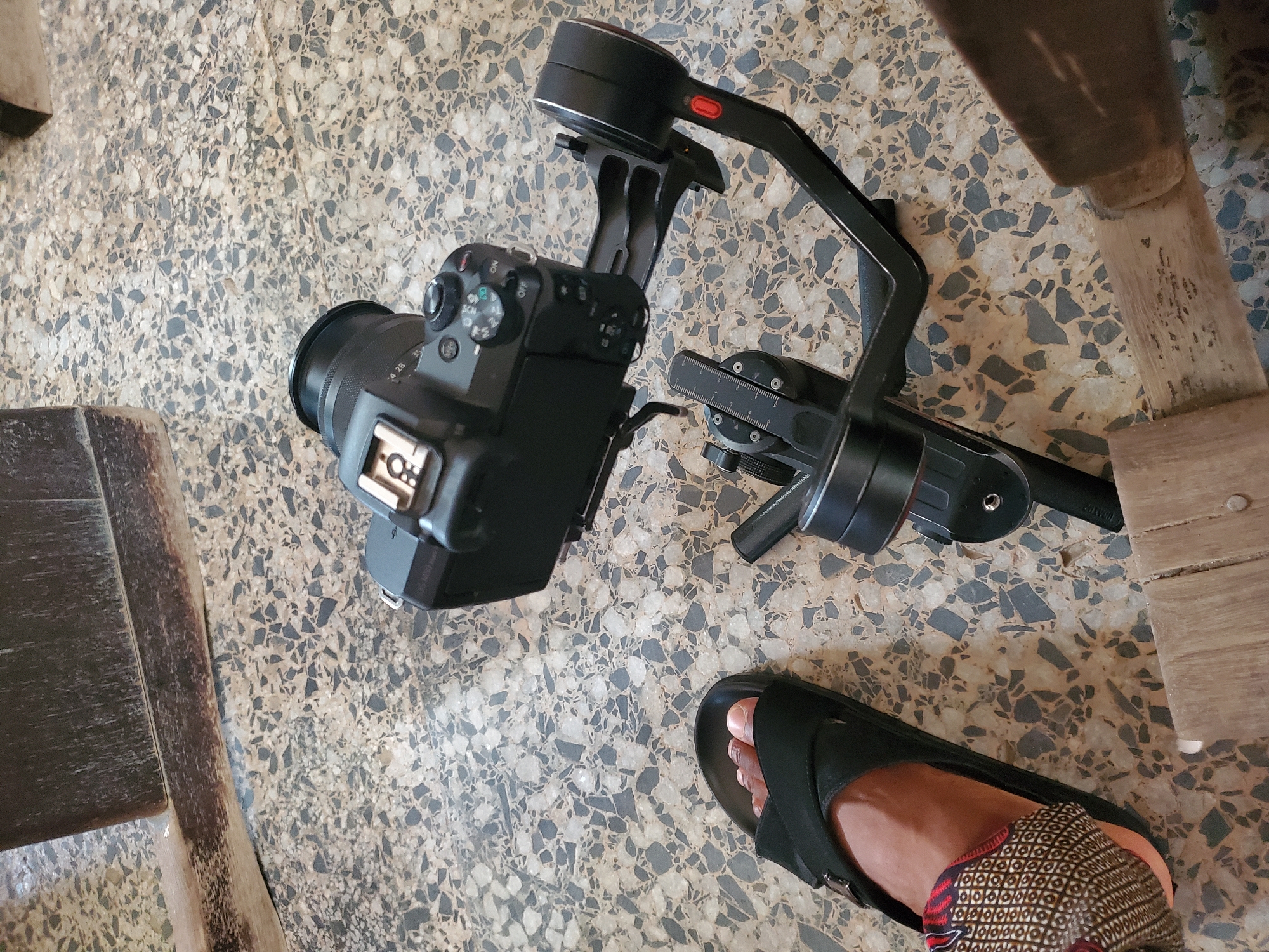 The image depicts a camera mounted on a gimbal lying on a terrazzo floor. A person wearing a sandal is partially visible in the lower right corner of the frame, with part of patterned trousers showing. Two wooden furniture legs are also visible on the edges of the frame. The location is undetermined, so that Fegge, Nigeria, is speculative.