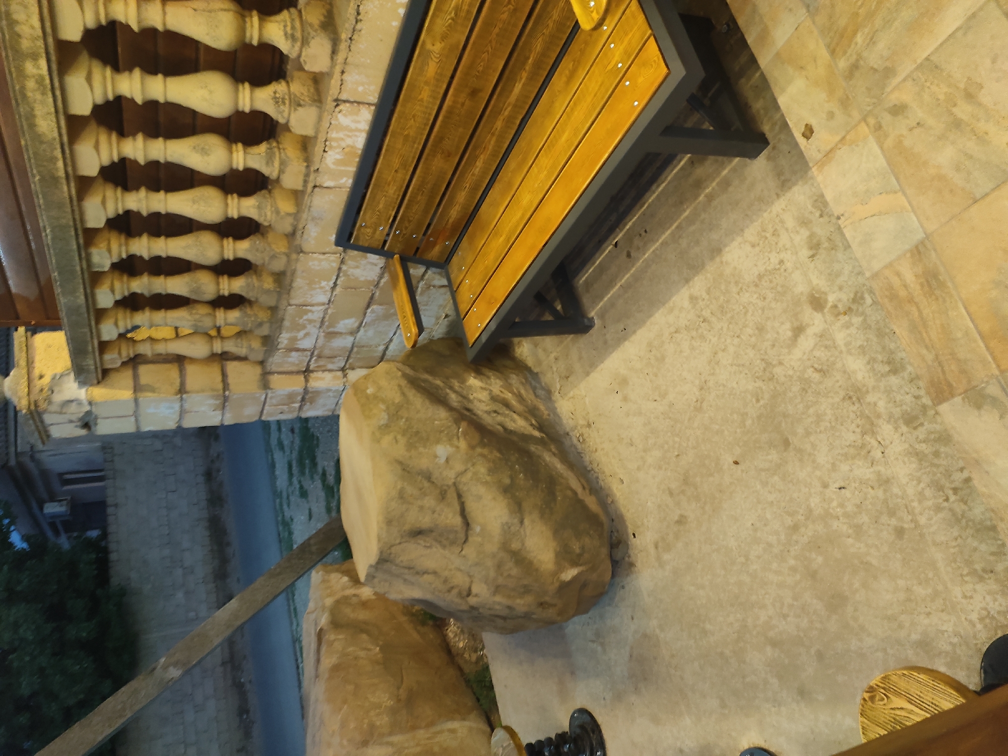 A wooden bench with a dark grey metal frame sits on a paved patio. The bench is oriented towards the right of the frame, with its back partially visible. To the left of the bench are two large, rough-textured, beige-colored rocks. One rock is in the foreground, and another is behind it. In the background, a low stone wall with decorative balusters runs horizontally across the upper portion of the image. Beyond the wall, a building with a dark roof and a light-colored facade is visible. A portion of a tree with dark green foliage can be seen to the left of the building. The ground in front of the bench and rocks is paved with light-colored concrete and stone tiles. The scene appears to be outdoors in an urban or suburban setting, possibly a park or a residential area in Leninkent, Russia.