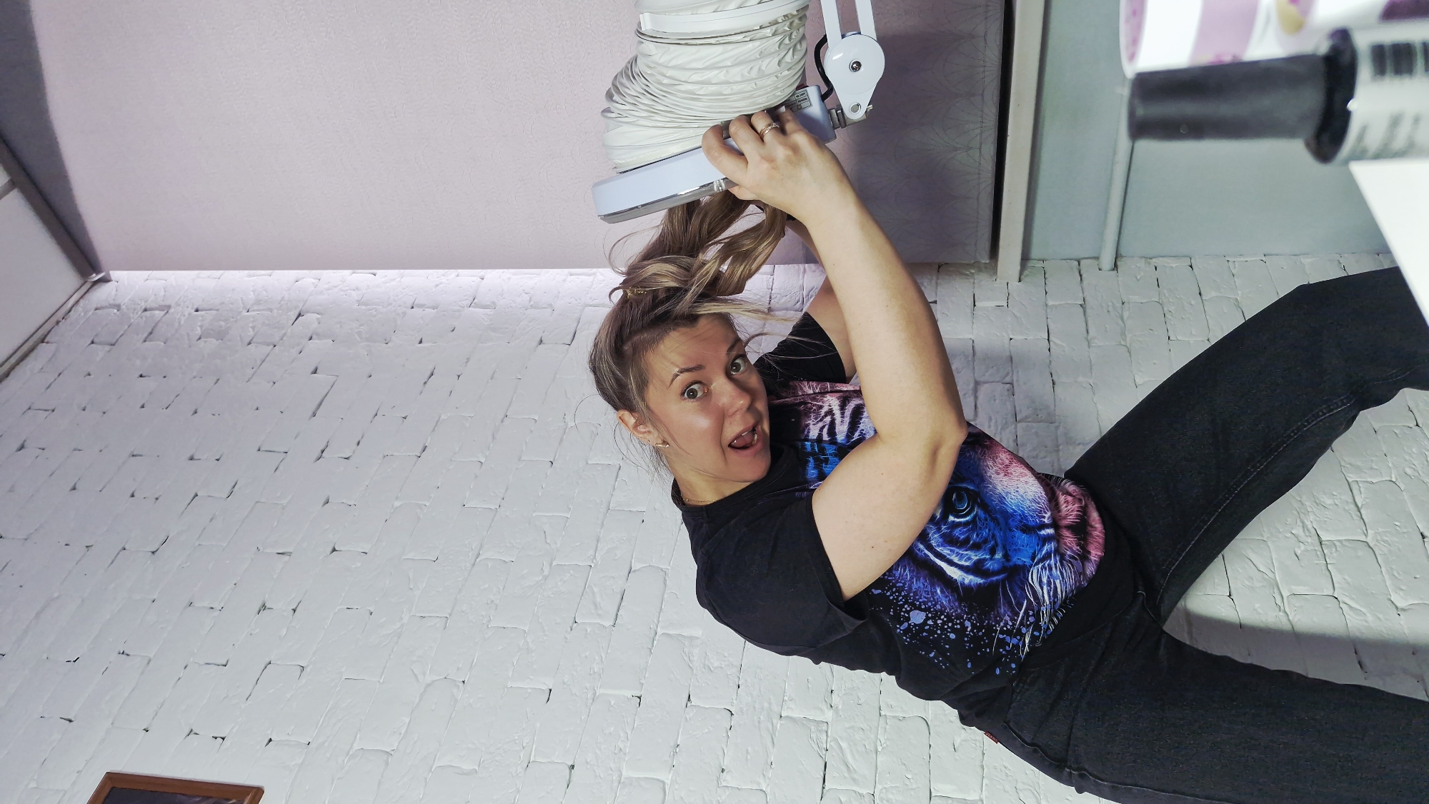 A young woman with blonde hair, styled in a casual, messy bun, is depicted in an inverted perspective, creating the illusion that she is hanging upside down. She is actively holding onto a white, accordion-like flexible duct or hose that appears to extend from the ceiling above her. Her facial expression is one of wide-eyed surprise and playful amusement, with her mouth slightly open.
She is dressed in a black t-shirt featuring a prominent, colorful graphic print of what appears to be a lion or tiger head with blue and purple tones, and dark-colored pants, likely jeans. The background consists of a white brick-patterned wall that covers the lower and central parts of the frame, contrasting with a plain, light-colored ceiling occupying the upper section. Elements resembling furniture or shelving units are partially visible on the periphery, to the left and right, and a small portion of a framed item can be seen in the bottom left corner.
The unconventional camera angle and inverted orientation of the image give it a dynamic and disorienting quality. The lighting suggests an indoor setting, possibly a room in Otchor, Russia. There is no visible text in the image.