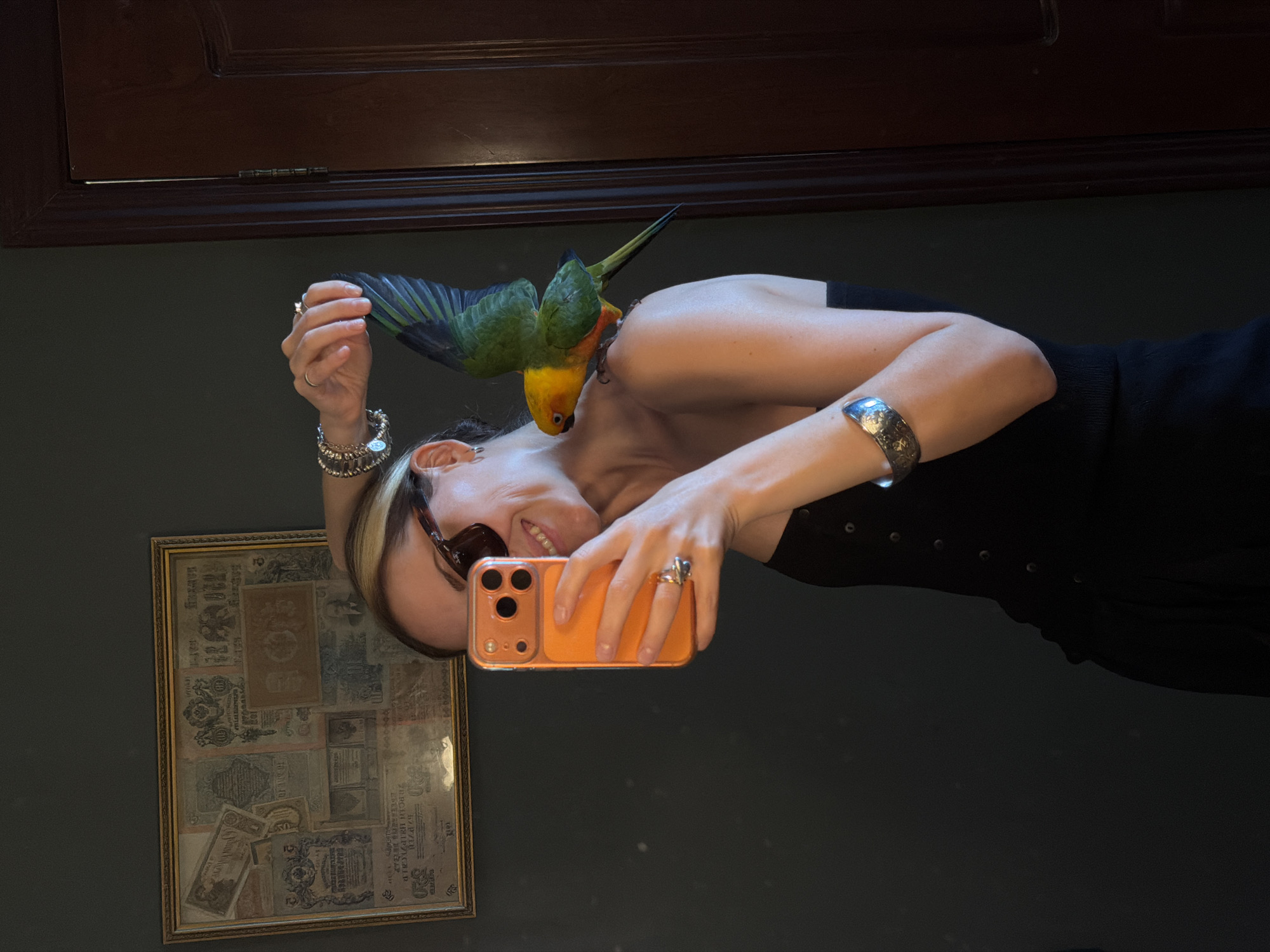 The image depicts an indoor scene in Khimki, Russia, featuring a fair-skinned individual in her 30s or 40s, captured via a mirror selfie. The person, wearing dark sunglasses and a black strapless top or dress, smiles towards the camera while holding an orange-cased smartphone in her right hand. Her left shoulder hosts a small parrot, displaying yellow and green plumage, with its left wing partially extended upwards. The individual's left wrist is adorned with multiple silver chain bracelets and a wide silver cuff on her forearm, while her right ring finger wears a prominent ring with a dark, possibly metallic, stone. In the background, a dark-colored wall is visible, on which a framed collage of various old banknotes or currency is mounted. To the far right, a dark wooden door frame and hinge are partially visible.