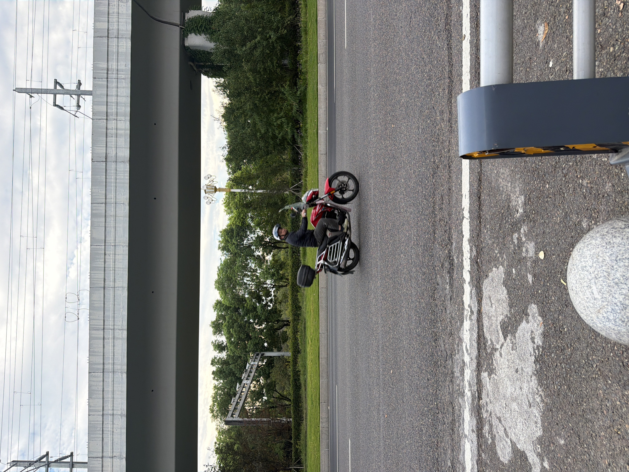 A person rides a red motorcycle with a black storage box on the back along a paved road in Jiangjin, China. The motorcycle has silver accents and a windshield. The rider wears a dark jacket and a helmet. Overhead, a concrete overpass supported by pillars stretches across the frame, with overhead power lines visible against a cloudy sky. The road is lined with green grass and trees on the left side. In the foreground, a portion of a metal barrier and a rounded stone object are visible.