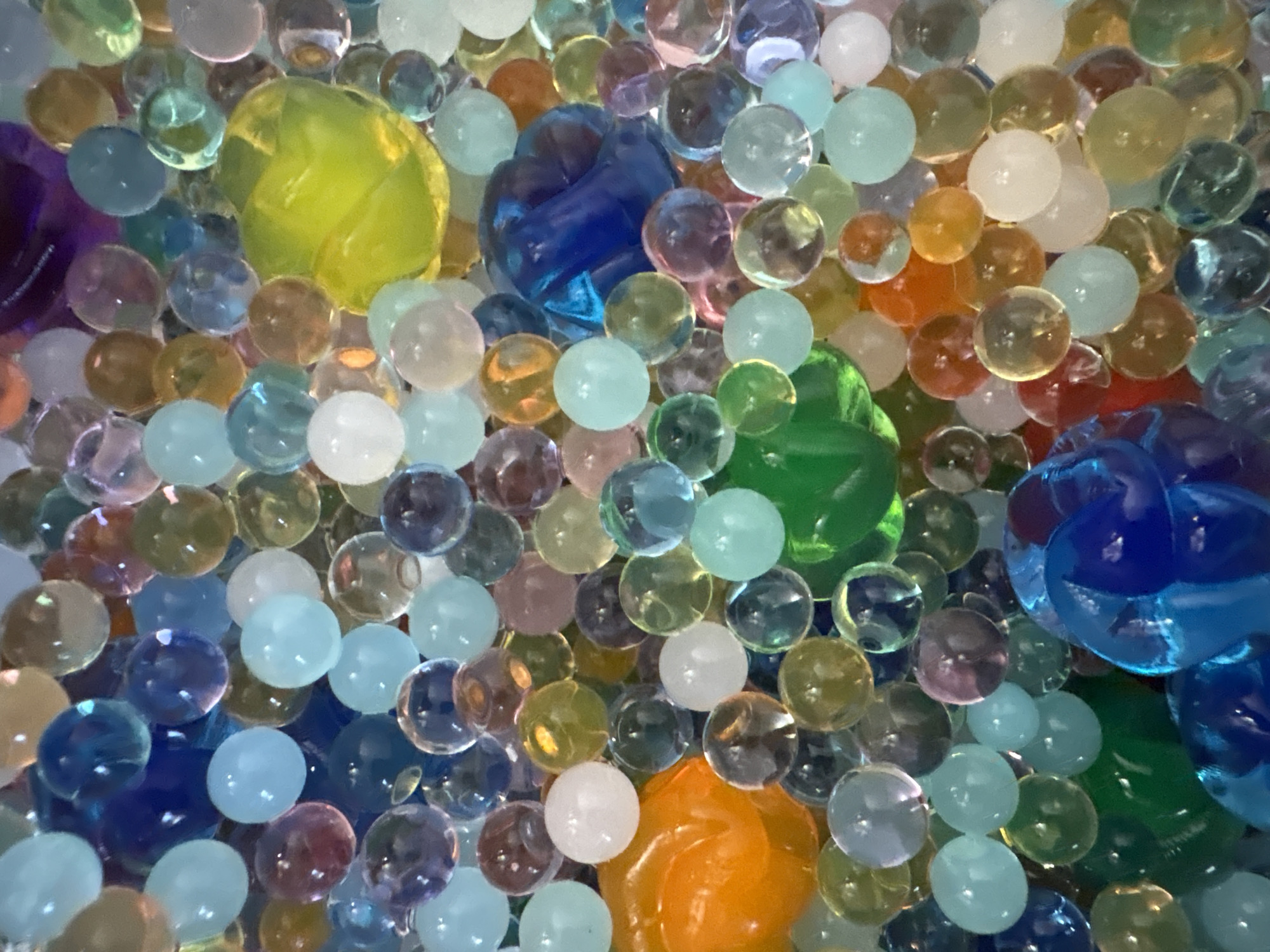 The image displays a close-up, overhead view of a pile of colorful, translucent, water beads. The beads vary in size and color, including shades of blue, green, yellow, orange, purple, and white. Some beads appear spherical, while others are more irregularly shaped. There are no discernible people, animals, or structures. The lighting suggests an indoor environment, but no specific location within a city can be determined.
