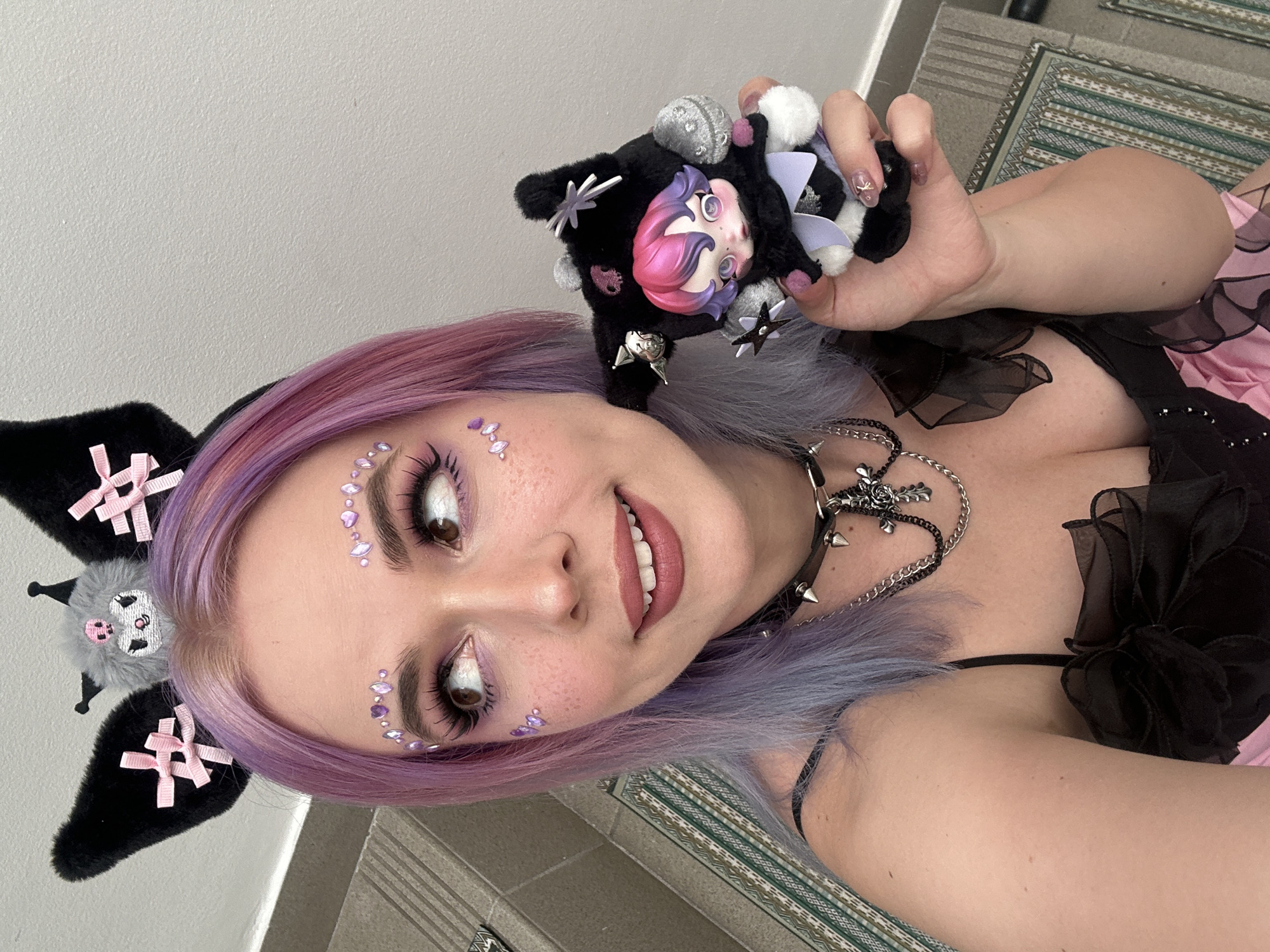 A medium shot features a fair-skinned woman holding a doll, angled to the left. The woman has violet-tinged hair, sparkling gems on her cheeks, and is smiling. She wears a black top with ruffles and a spiked choker, along with multiple silver necklaces. On her head, she has a black and grey headpiece with pink bows.
She is holding a black, fuzzy doll with accessories like silver stars and spikes. The doll has a pink-haired figurine attached to it.
The background consists of a white wall, a patterned rug, and a glimpse of the woman's pink and black outfit.
There is no information to indicate the location is Zagreb, Croatia.