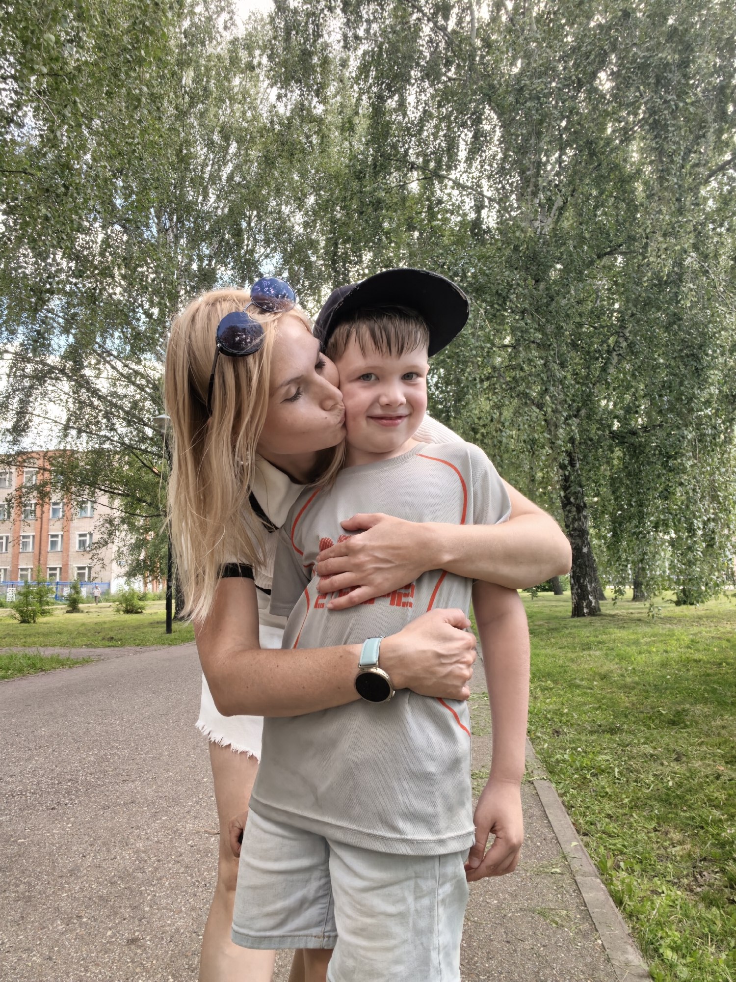 In Sterlitamak, Russia, a blonde woman wearing sunglasses on her head, a white top, and a white skirt kisses a young boy on the cheek. The boy is wearing a grey t-shirt, grey shorts, and a black baseball cap. They are standing outdoors on a paved path in a park with green grass and trees. In the background, there is a building with multiple windows and balconies, possibly an apartment complex. The overall atmosphere appears to be a casual outdoor setting on a pleasant day.