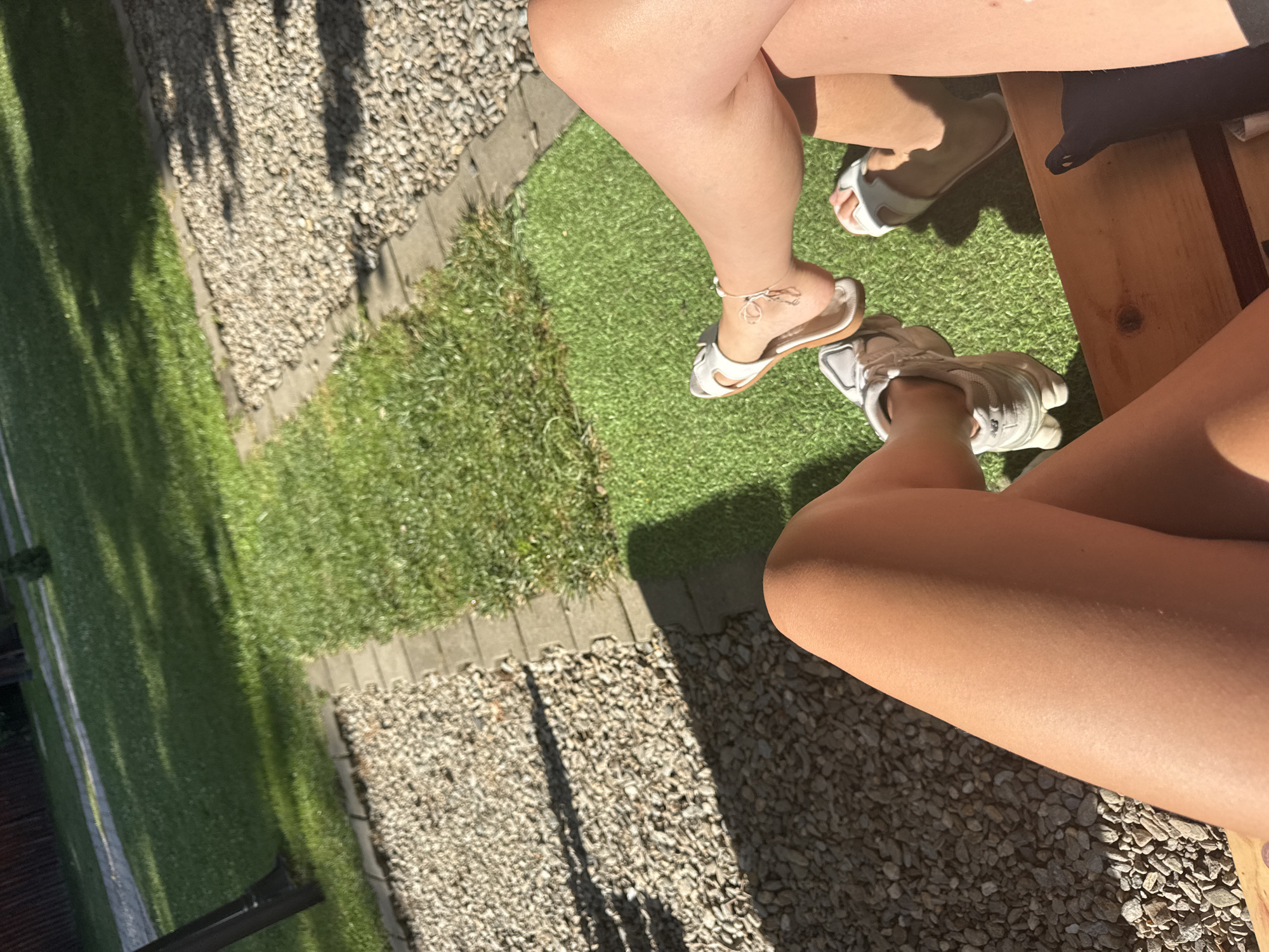 The image depicts two individuals sitting outdoors, with their legs visible. The person on the left is wearing white sneakers and is partially obscured by their crossed leg. Their skin appears tanned. The person on the right is wearing white slides and has a small tattoo on their ankle. Their skin is also tanned. They are sitting on a wooden surface, possibly a bench, in front of a patch of artificial turf. The background shows a gravel path bordered by grass and some trees, suggesting a garden or yard setting. The lighting indicates it is daytime, with shadows cast by foliage. The specific location of Cârțișoara, Romania is not visually ascertainable from the provided image alone.