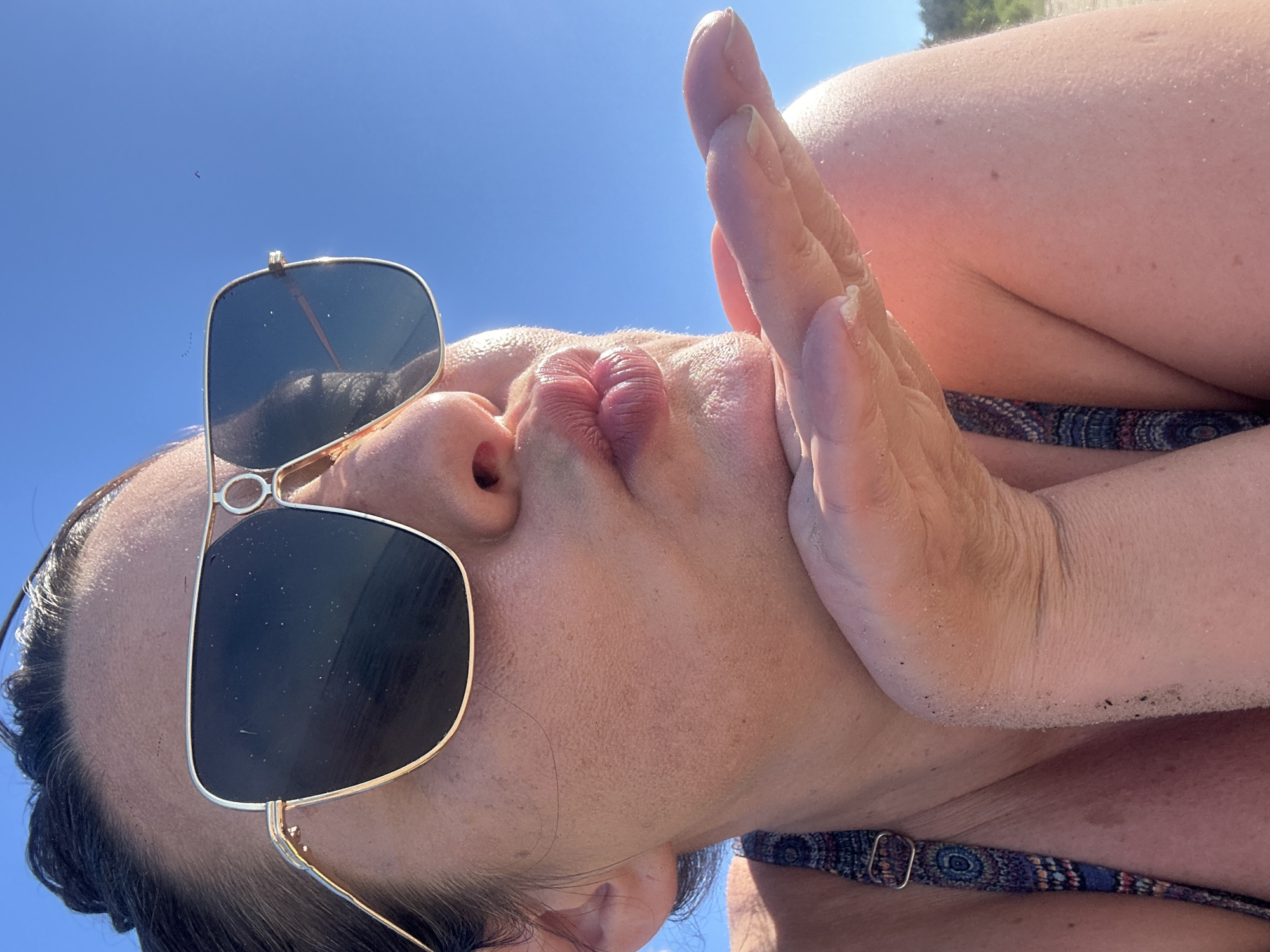 A close-up, eye-level, outdoor, medium shot captures a woman from the shoulders up, against a clear blue sky. She is wearing large, gold-rimmed sunglasses and a patterned bikini top. Her left hand is extended towards the camera, palm facing outwards, as if blowing a kiss. Her lips are pursed, and her skin is slightly flushed. The background appears to be a beach setting, with subtle hints of sand on her hand and shoulder, and what looks like foliage in the far distance, suggesting a location in Jūrmala, Latvia. The sunlight highlights the texture of her skin and the sand.