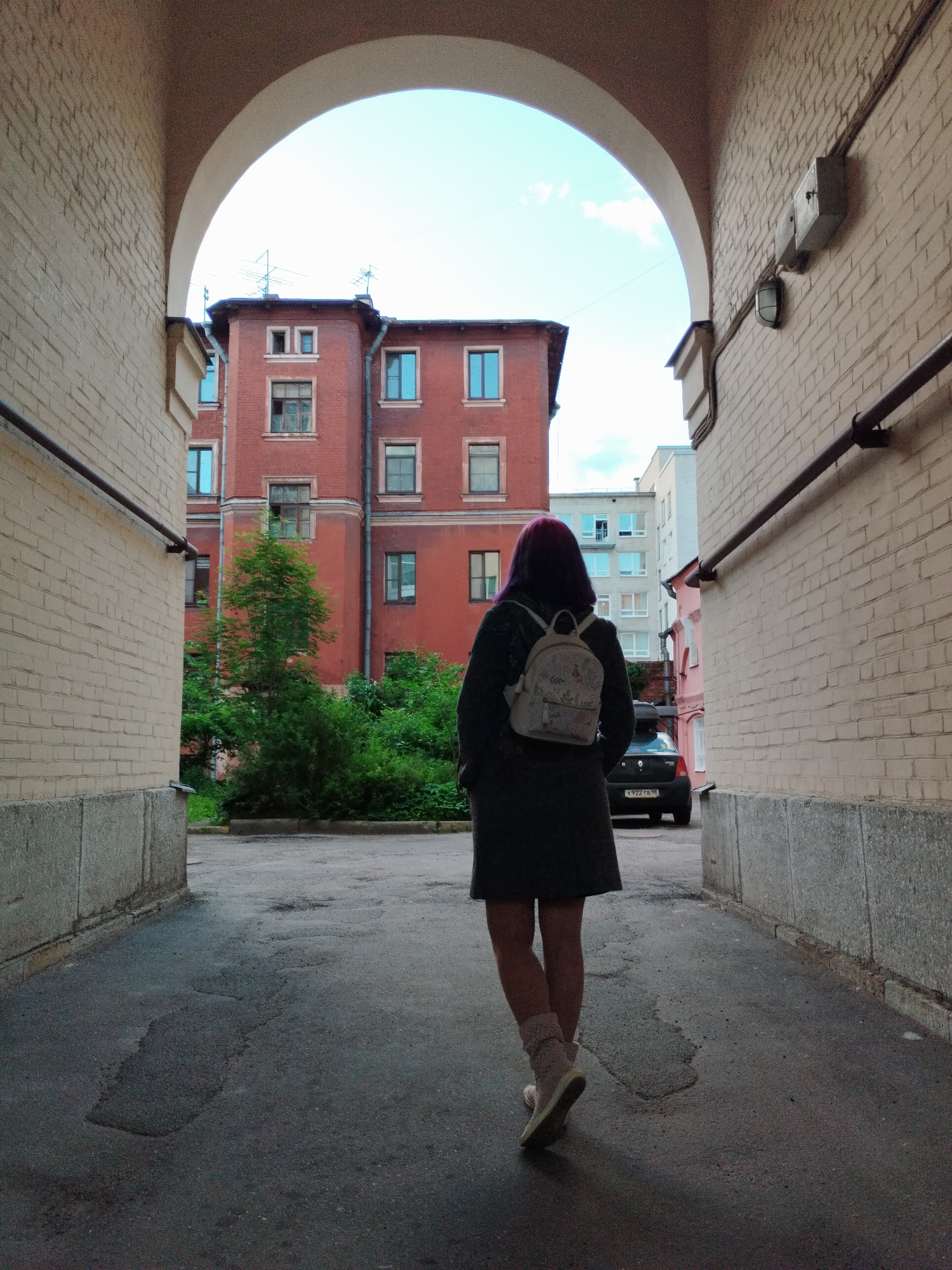 An overcast day in a Saint Petersburg, Russia, courtyard, viewed from under an archway. A person with dark hair, wearing a dark coat and a light-colored backpack, walks away from the camera on a paved pathway. The pathway is uneven with patches of darker asphalt. To the left, a beige brick wall forms the side of the archway, with a grey concrete foundation. To the right, another beige brick wall continues, with electrical conduits and a wall-mounted box. Beyond the archway, a red brick building with multiple windows is visible, along with green trees and bushes. Further in the background, lighter-colored apartment buildings can be seen under a pale sky. A dark car with a license plate is parked in the distance.