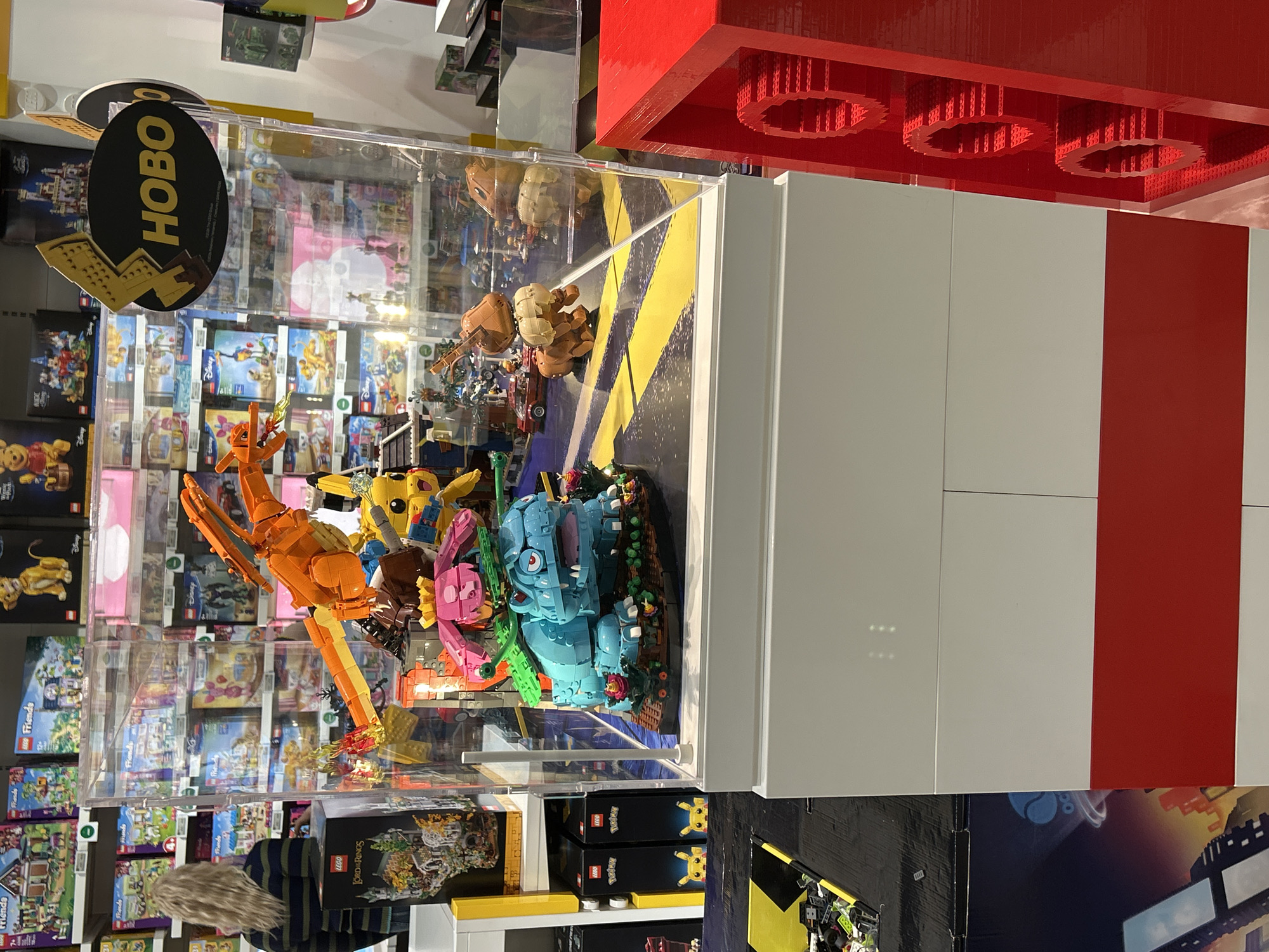 The image shows a display case filled with Lego models of cartoon characters, including Pokemon. The display is inside a store, with shelves of Lego sets visible in the background. A sign reading "HOBO" is attached to the case. The location is the Hobo toy store in Sofia, Bulgaria. The display case is built on a platform made of large Lego bricks in white and red. A black and yellow road marking design is on the platform under the models.