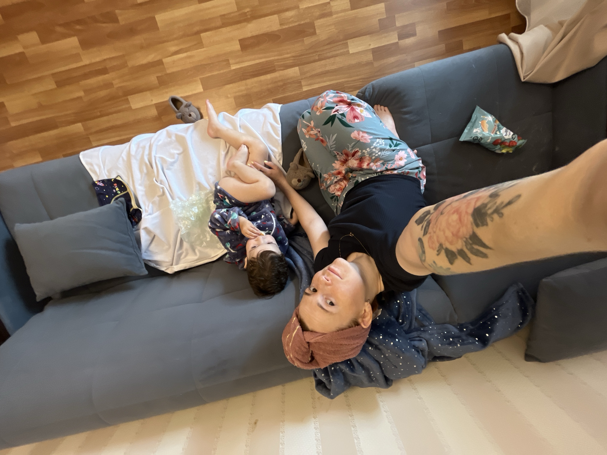 An indoor scene in Chaikovsky, Russia, captured from an overhead selfie perspective. An adult female and a young child are seated on a dark grey upholstered sofa. The adult, positioned on the left, looks directly at the camera with a faint smile. She has light skin, a reddish-brown hair towel wrapped turban-style with a subtle gold stripe, a black ribbed top, and floral patterned wide-leg pants (grey-blue with pink, orange, and white flowers and green leaves). A prominent floral tattoo in red and green is visible on her right forearm, which extends into the frame from the left, holding the camera. A thin gold-colored necklace is visible. Her bare feet rest on the sofa. To her right, a young child, appearing to be a toddler, sits facing slightly away from the camera, occupied with a small white object held to its mouth. The child wears dark blue long-sleeved pajamas patterned with white celestial objects. Bare feet are visible. A light blue blanket with white polka dots is draped over the back of the sofa on the left. A small teal snack bag with red fruit shapes is on the sofa cushion near the adult's left leg. A white sheet or blanket is partially draped over the sofa between the child and the backrest. Two grey animal-shaped soft slippers are on the sofa near the individuals' feet. The floor is light brown wood laminate, and the wall in the background is light-colored with subtle vertical stripes.