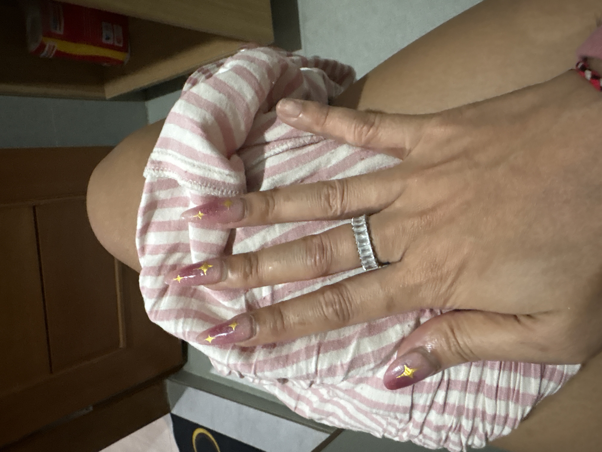 The image presents a close-up view of a person's hand resting on a pink and white striped garment. The hand has a medium skin tone and features long, pointed, manicured nails with a pink and purple gradient design, adorned with small gold star decals. A prominent silver-toned eternity band ring, set with rectangular clear stones, is worn on the ring finger. A portion of the person's upper arm or thigh is visible in the upper right, and a red, black, and white patterned bracelet is faintly visible on the wrist.
The striped garment, possibly shorts or pajamas, has a gathered elastic waist and a soft texture. In the background, to the upper left, a red and yellow rectangular carton is placed on a light wooden shelf. The letters "BB" are clearly visible on the side of the carton. Below the shelf, darker wooden furniture, possibly a cabinet or paneling, is visible. At the very bottom right, a sliver of a black mat or rug with a yellow circular design can be seen. The overall setting appears to be indoors, suggesting a casual, personal space like a bedroom or living area. The image captures a static pose, highlighting the hand and its adornments.