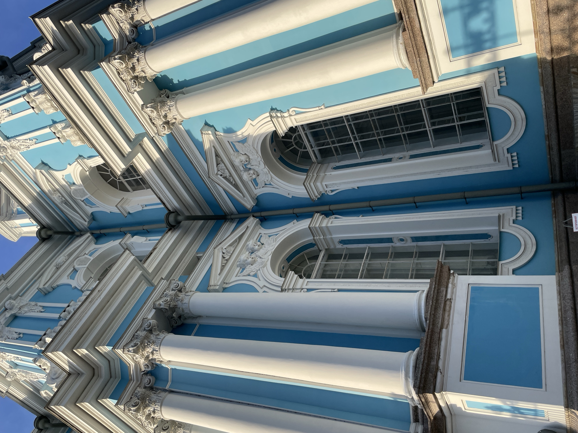 This image displays a low-angle, upward-looking view of the Smolny Cathedral in St. Petersburg, Russia, focusing on a corner of its exterior facade. The building is characterized by its distinct Russian Baroque architectural style, featuring a vibrant light blue color on its walls contrasted with white architectural elements. White Corinthian columns with elaborate capitals are prominently positioned against the blue walls, casting distinct shadows. Ornate white stucco decorations adorn the cornices, window frames, and upper sections, including intricate scrollwork, pediments, and sculpted cherubic figures (putti) above some windows. Tall, multi-paned arched windows with white frames are visible, reflecting ambient light. The scene is brightly lit by natural sunlight, creating strong contrasts and shadows that accentuate the three-dimensional relief of the architectural details. A section of a grey foundation or base is visible at the bottom, and a hint of a Russian cautionary sign ("ОСТОРОЖНО!") is visible in one of the lower cropped sections. No people or active elements are present in the image.