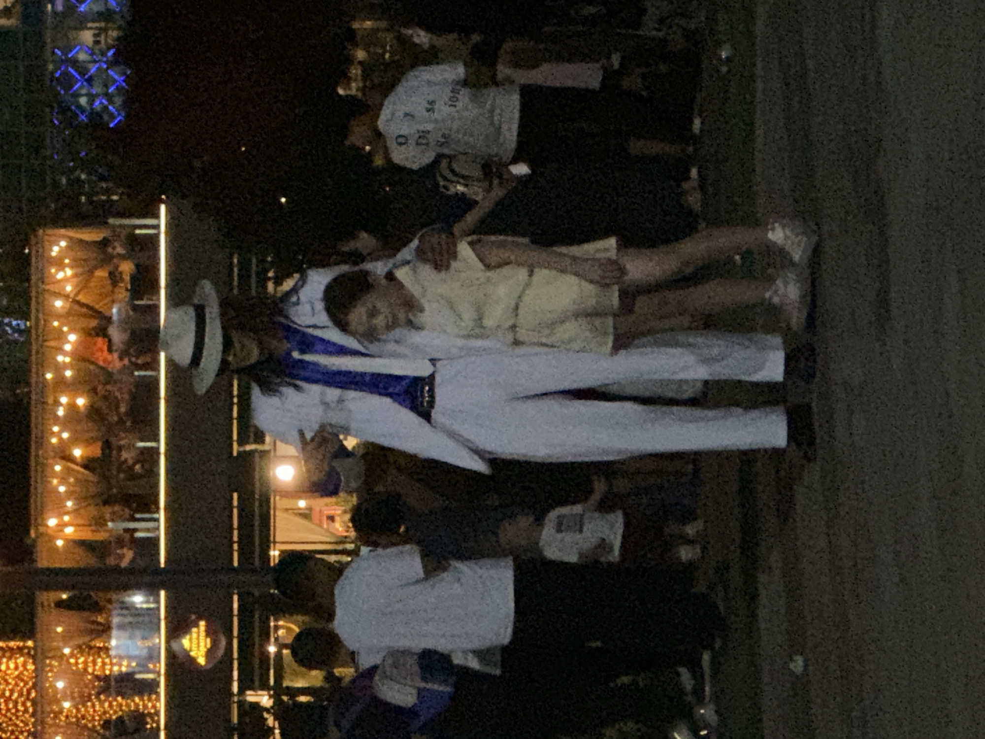 The image captures a nighttime scene at an outdoor venue, possibly a restaurant or entertainment establishment, in Tashkent, Uzbekistan. In the foreground, a person dressed in a white suit, white fedora hat, and blue shirt, reminiscent of Michael Jackson's attire, stands with a young girl wearing a yellow outfit. The person in the white suit has their arm around the girl's shoulder, suggesting an interaction between them. Several other people are visible in the background, appearing as silhouettes or less distinct figures, some wearing casual attire like white t-shirts. The environment is illuminated by decorative string lights and architectural lighting, creating a warm and festive atmosphere. A sign with a logo and text, "Arrows & Sparrows," is visible on a building in the background, indicating the establishment's name. The ground appears to be paved or asphalted. The overall impression is of a lively gathering in an urban setting at night.