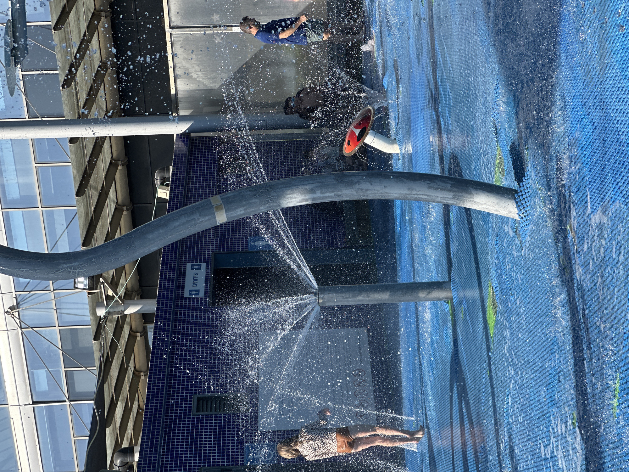 This is a sunny, outdoor image of a water play area in Hod HaSharon, Israel. The ground is covered in a blue, textured surface with water spraying from several points. In the foreground, a large grey pipe curves upwards, and to its right, a red and white mushroom-shaped water feature is partially visible.
On the left side of the image, a young person, appearing to be a child or pre-teen, is standing with their back to the camera, wearing a leopard-print top and brown shorts. They are surrounded by water spray. Further back, a building with a blue tiled facade is visible. Above the tiles, a sign with Hebrew text and restroom symbols is present.
To the right of the building, a man in a blue t-shirt and dark shorts is walking away from the camera. He is also in the water spray. Behind the mushroom-shaped feature, a dark-haired figure, possibly a child, is visible, partially obscured by water. The background features a modern building with large glass windows and a covered walkway structure. The overall scene depicts a public recreational space on a bright day.
