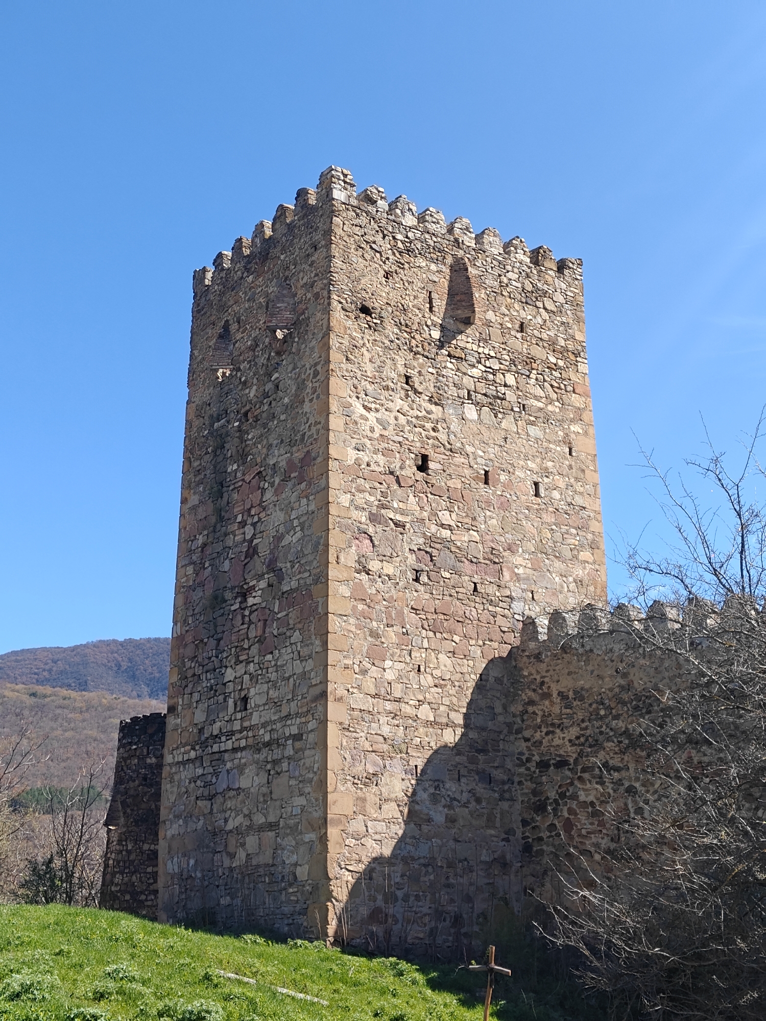 The image shows a tall, stone tower, likely part of a fortification or castle, against a clear blue sky. The tower is constructed of various sized stones, with crenellations at the top. There are small openings or windows in the tower walls. To the right of the tower is a stone wall, partially obscured by a leafless tree. In the background, there's a view of rolling hills or mountains. In the foreground, there is green grass. The location is in the city of Unknown, Unknown.