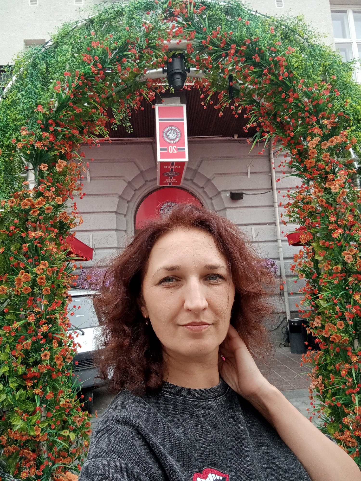 A woman with auburn, curly hair stands in front of an archway decorated with artificial greenery and small red and orange flowers. She is wearing a dark gray sweatshirt with a graphic of lips on the front. Her right hand is touching her neck. Behind her, a building with a gray facade and arched doorway is visible. A red sign with white text hangs above the doorway. The image appears to be taken outdoors in a city environment. The specific location in Kuopio, Finland is not discernible from the visual information provided.