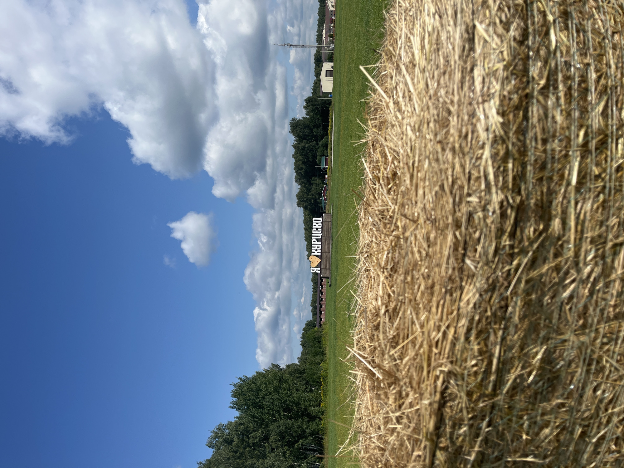 In Akhlebaikha (Kovriginskii S/S), Russia, a close-up shot of a bale of hay dominates the foreground, its dry, golden strands filling the bottom half of the frame. Beyond the hay, a vibrant green field stretches towards a line of trees in the mid-ground. A sign with large white letters and a yellow heart, reading "Я КУРИЦКО" (Ya Kuricko), is visible in the center of the field. In the background, several buildings, including what appears to be a warehouse and a tall tower, are situated along the tree line under a bright blue sky dotted with fluffy white clouds.